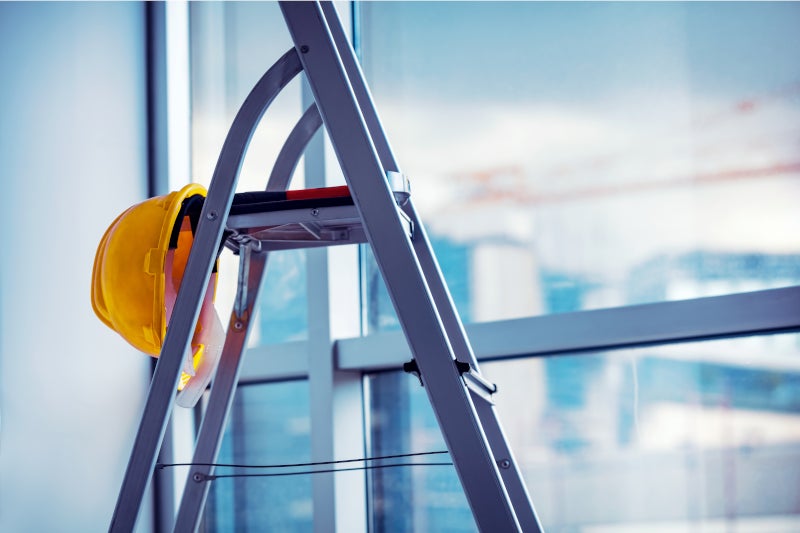 Leiter mit gelbem Helm vor Fensterfront, im Hintergrund unscharf Baustelle zu erkennen
