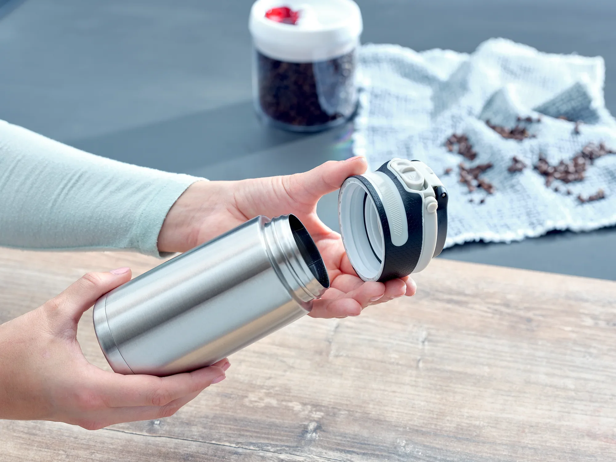 Izolowany kubek Flip 350ml w kolorze srebrnym jest otwarty. Osoba trzyma pokrywkę w jednej ręce, a kubek w drugiej. W tle widać ziarna kawy.