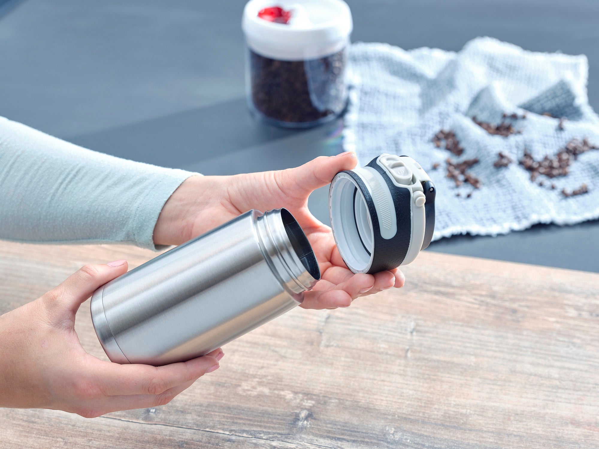 Geïsoleerde mok Flip 350ml in zilver wordt geopend. Een persoon houdt het deksel in de ene hand en de mok in de andere. Koffiebonen zijn te zien op de achtergrond.