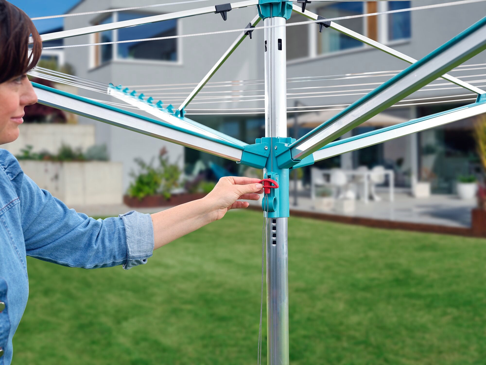 Eine Person bedient den Wäscheschirm Linomatic 500 Easy im Garten. Der Schirm ist geöffnet und zeigt seine robuste Konstruktion. Im Hintergrund ist ein modernes Haus zu sehen.