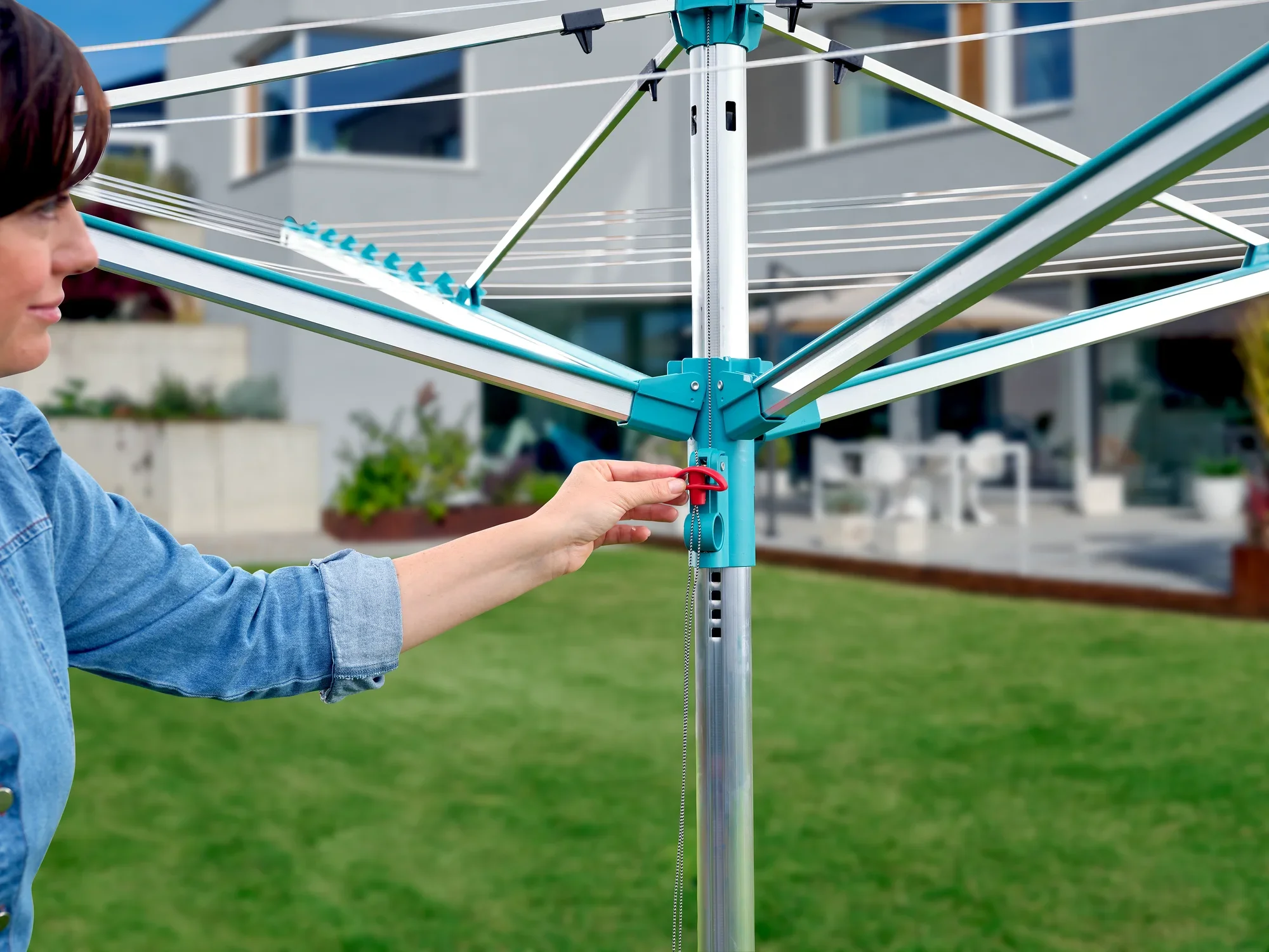 Osoba obsługuje parasol do prania Linomatic 500 Easy w ogrodzie. Parasol jest otwarty i pokazuje swoją solidną konstrukcję. W tle widać nowoczesny dom.