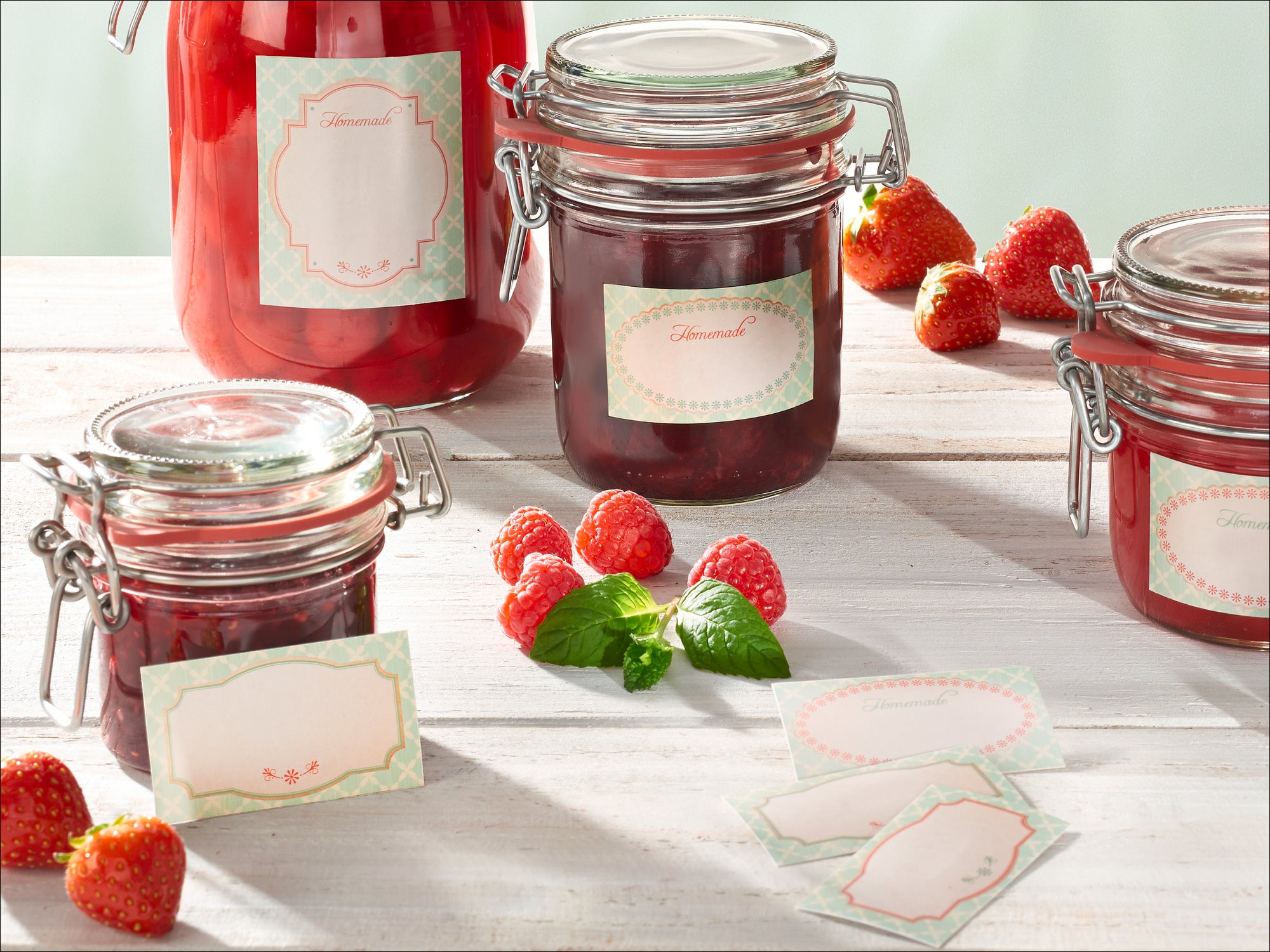 Verschiedene Einmachgläser mit roten Früchten und dekorativen Etiketten auf einem weißen Tisch. Frische Erdbeeren und Himbeeren liegen daneben, umgeben von Minzblättern.