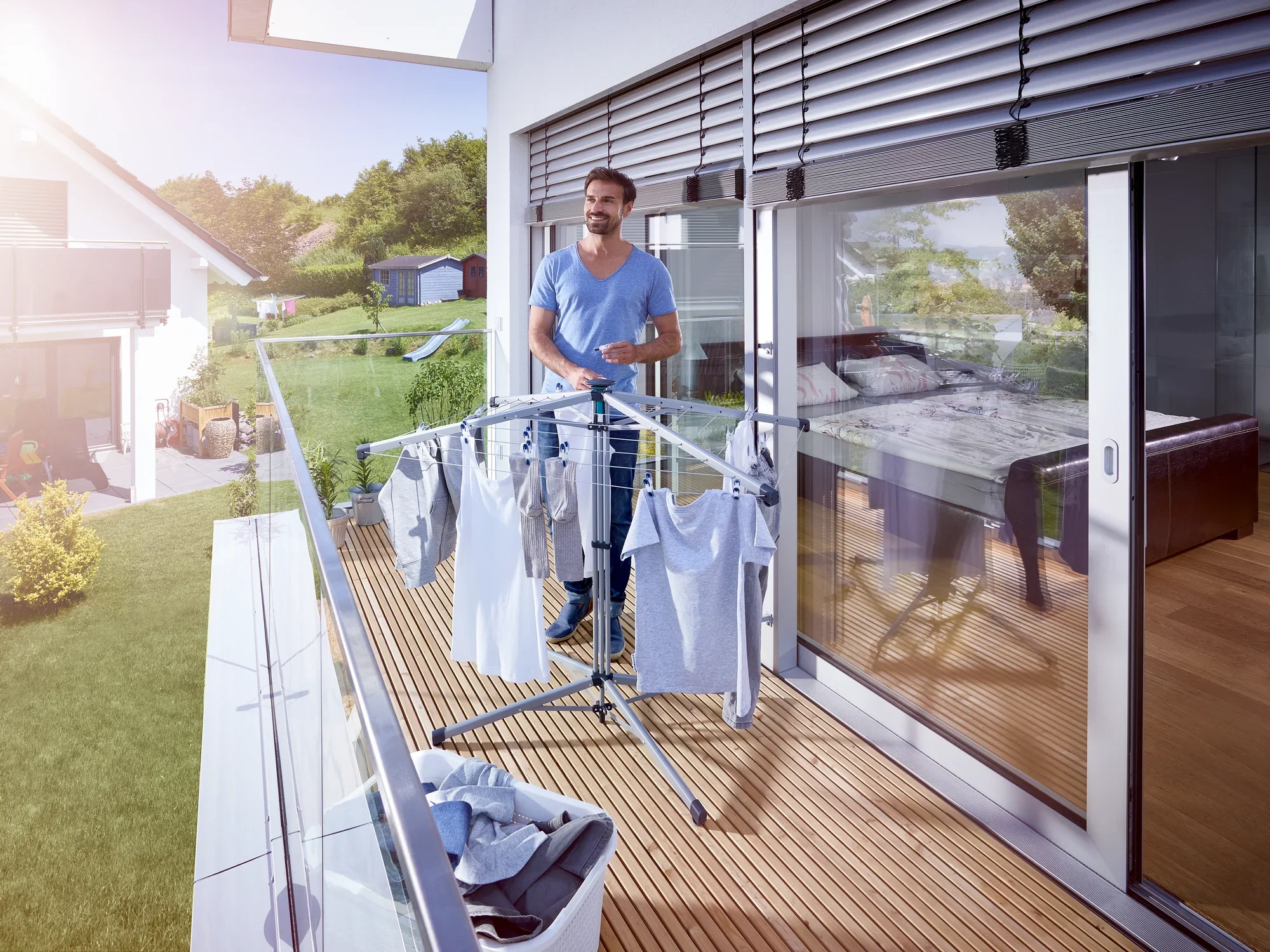 Un hombre está de pie en un balcón soleado y utiliza el tendedero de pie Lino Pop-Up para secar la ropa recién lavada. Al fondo se ve una casa moderna.