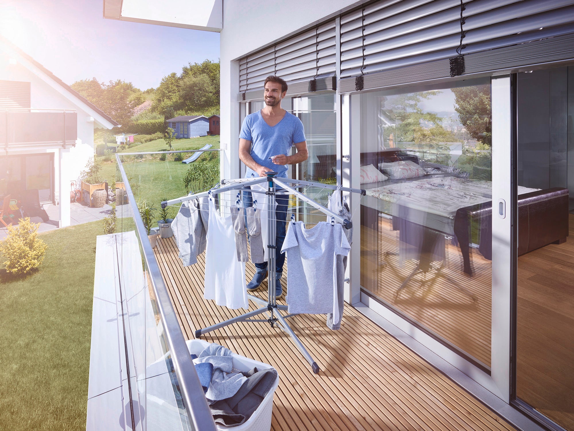 Ein Mann steht auf einem sonnigen Balkon und benutzt die Standwäschespinne Lino Pop-Up, um frisch gewaschene Kleidung zu trocknen. Im Hintergrund ist ein modernes Haus zu sehen.