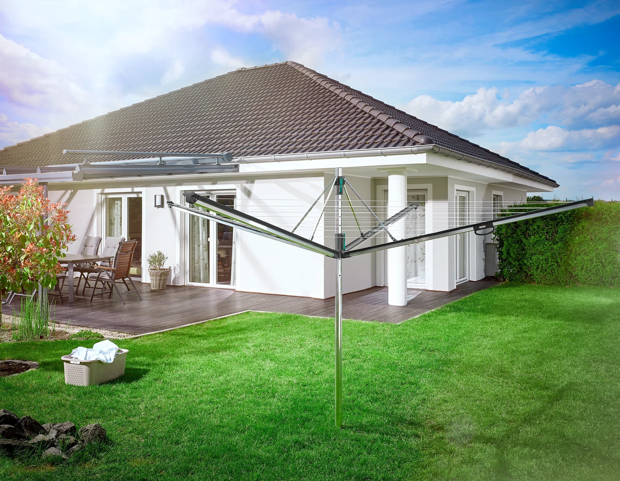 Un séchoir parapluie Leifheit Linomatic 500 Plus se trouve dans le jardin d'une maison moderne. La pelouse est bien entretenue et, à l'arrière-plan, on aperçoit une terrasse avec des meubles de jardin.
