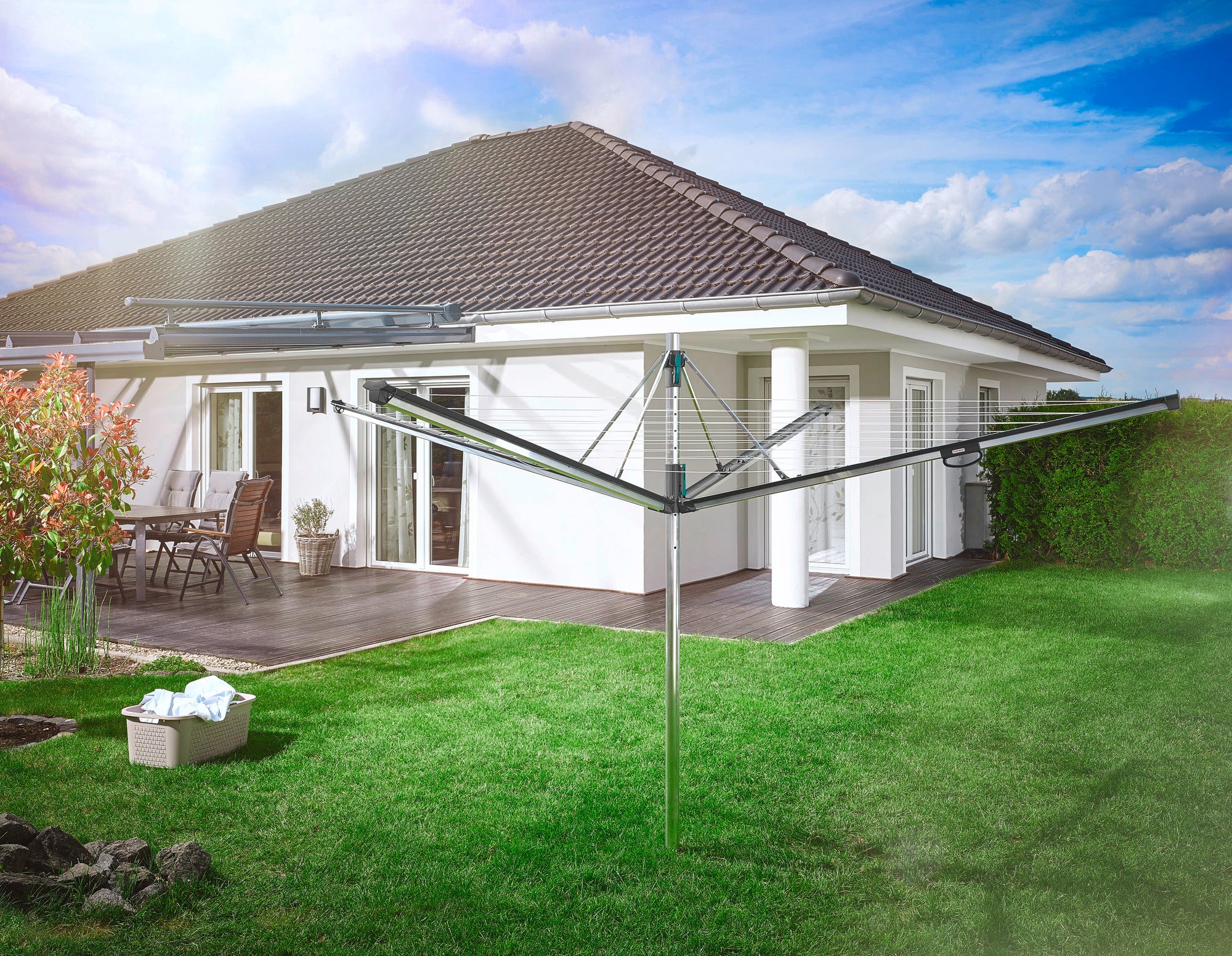 A Leifheit Linomatic 500 Plus rotary clothes dryer stands in the garden of a modern house. The lawn is well-kept and a patio with garden furniture can be seen in the background.