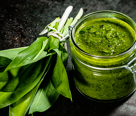 Ein Glas mit gruenem Baerlauchpesto neben einem Bund frischem Baerlauch auf dunklem Untergrund, die Blaetter sind leuchtend gruen.