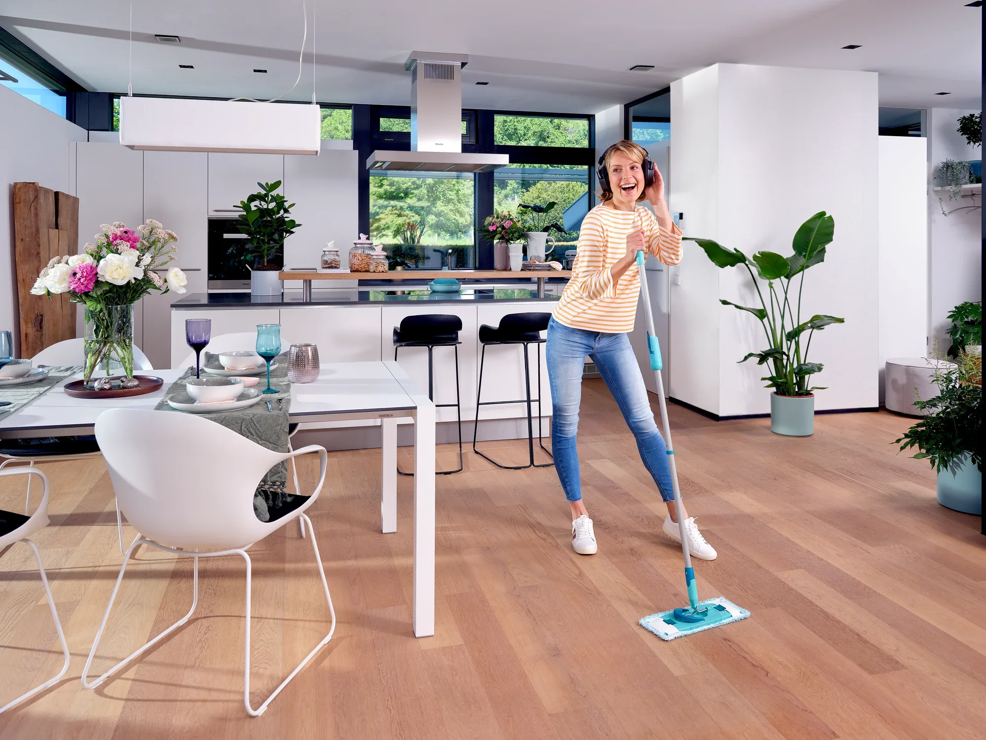 Une femme passe la serpillière avec le CLEAN TWIST M Ergo dans une cuisine moderne et lumineuse. Le sol est en bois et des plantes et des fleurs décorent la pièce. Elle sourit et semble prendre plaisir à faire le ménage.