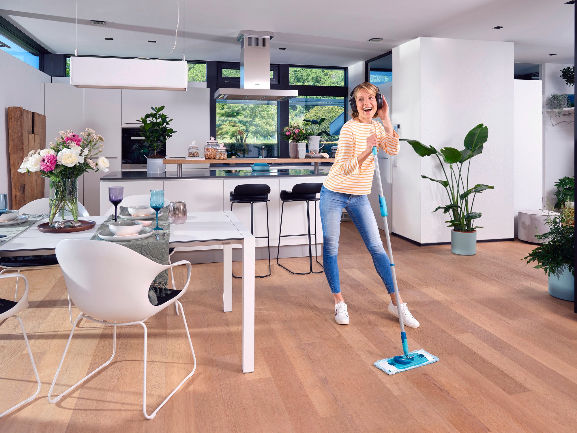 Une femme passe la serpillière avec le CLEAN TWIST M Ergo dans une cuisine moderne et lumineuse. Le sol est en bois et des plantes et des fleurs décorent la pièce. Elle sourit et semble prendre plaisir à faire le ménage.