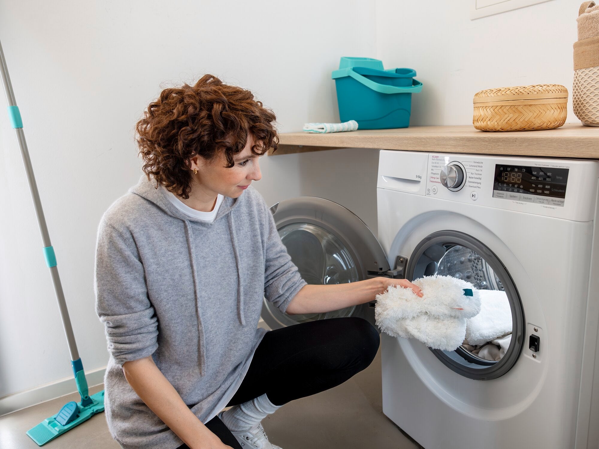 Een vrouw stopt de SUPERDUSTER XL plumeau in de wasmachine. De plumeau is groot en pluizig, ideaal voor moeilijk bereikbare plaatsen. Op de achtergrond zijn schoonmaakspullen te zien.