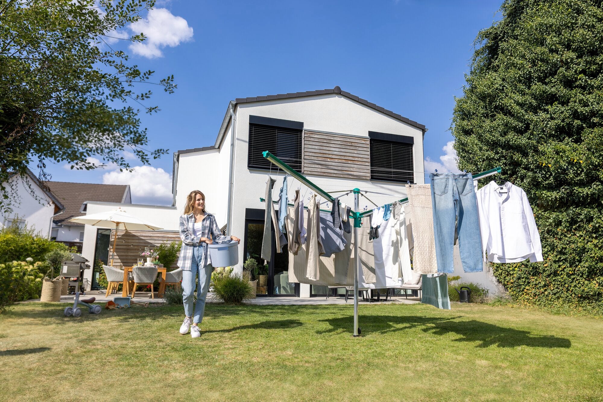 Eine Frau steht im Garten vor einem modernen Haus und hängt Wäsche an die Leifheit Wäschespinne Linomatic 500 Deluxe. Die Wäschespinne ist mit verschiedenen Kleidungsstücken behängt.