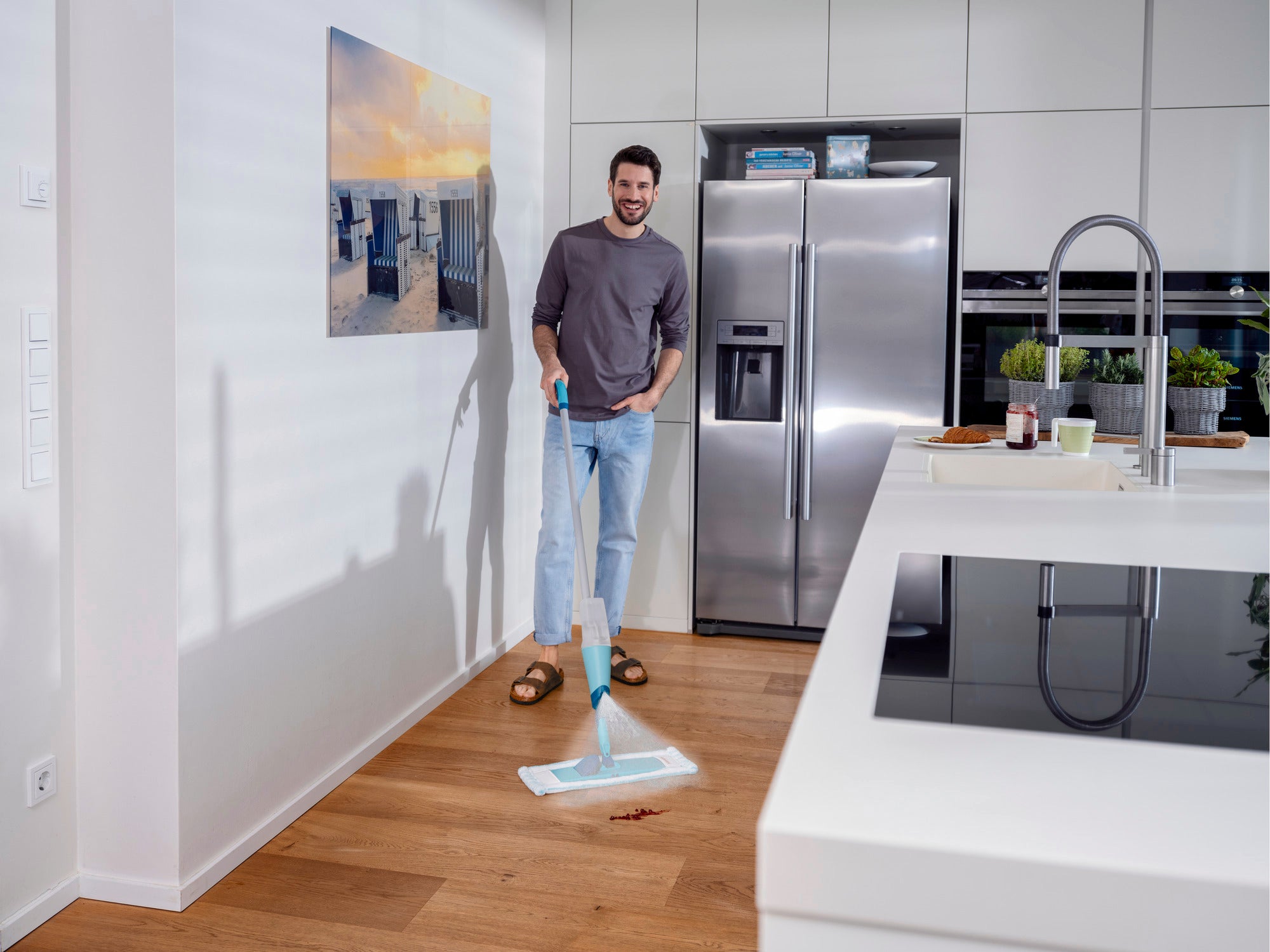 Un uomo è in piedi in una cucina moderna e utilizza il lavapavimenti a spruzzo Easy Spray XL per pulire il pavimento in legno. Il lavapavimenti ha un design blu ed è ideale per una pulizia rapida.