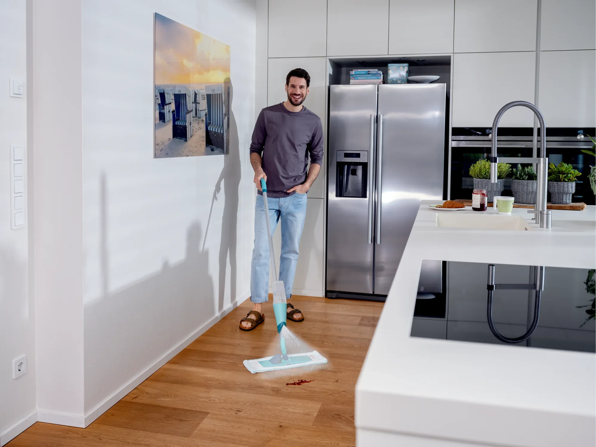 Ein Mann steht in einer modernen Küche und benutzt den Sprühwischer Easy Spray XL, um den Holzboden zu reinigen. Der Wischer hat ein blaues Design und ist ideal für die schnelle Reinigung.
