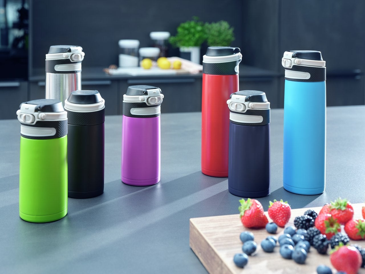 Verschiedene bunte Thermosflaschen stehen auf einem Kuechentisch neben einem Holzbrett mit frischen Erdbeeren, Brombeeren und Blaubeeren.