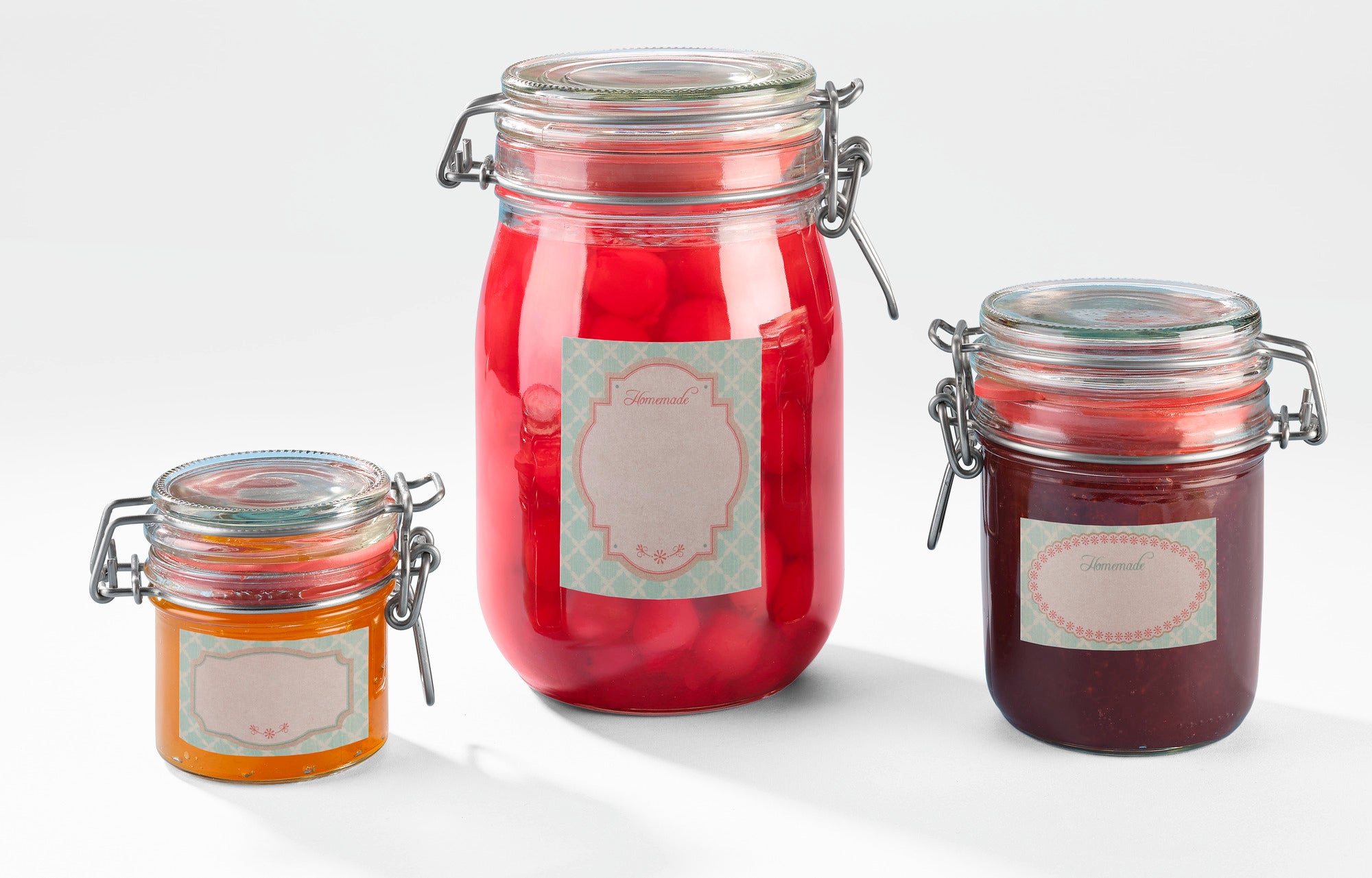 Drei Einmachgläser mit dekorativen Pastell-Etiketten. Das größte Glas enthält rote Kirschen, das mittlere Glas hat eine dunkle Marmelade und das kleinste Glas enthält eine orangefarbene Sauce.