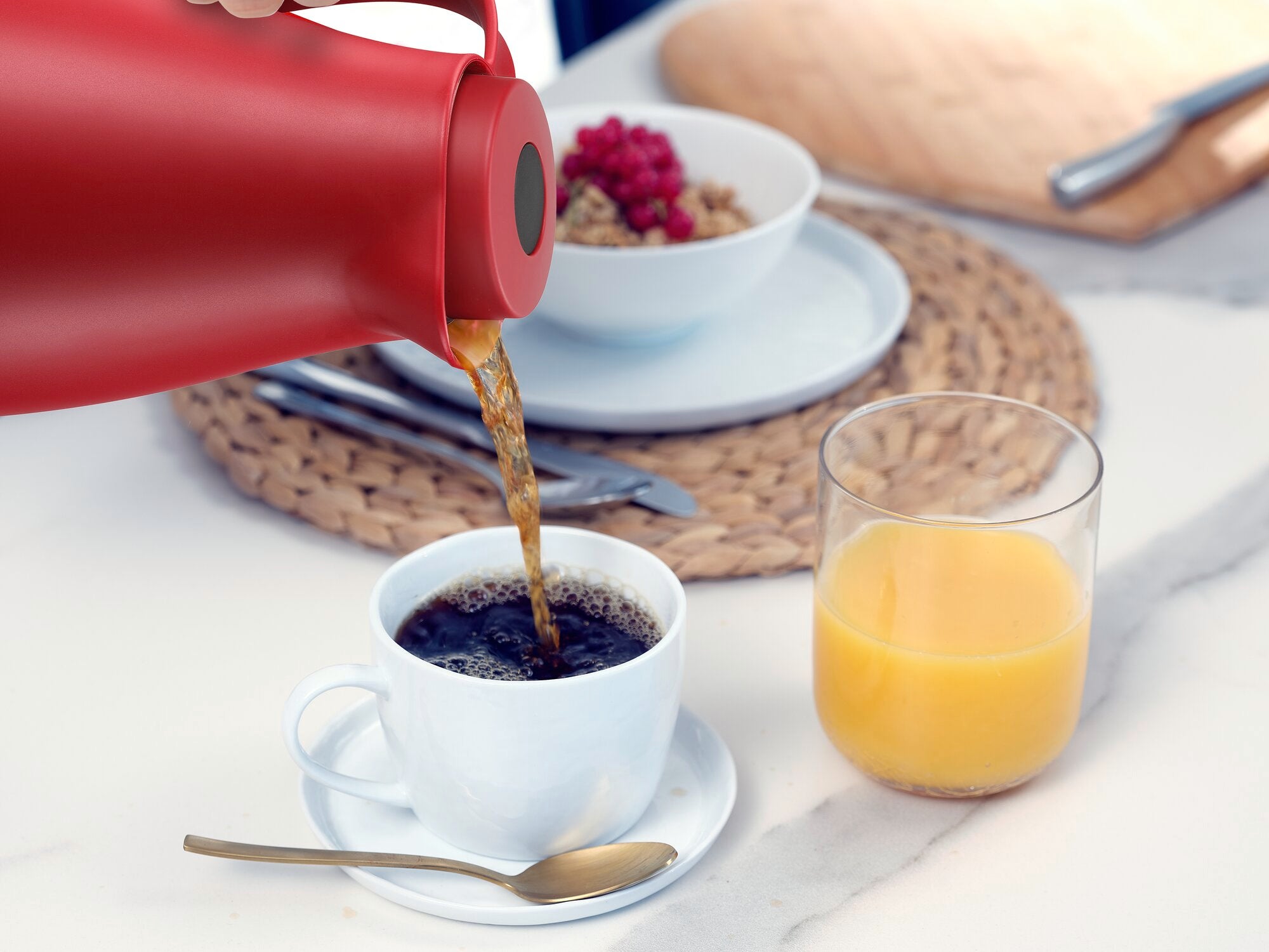 Een rode isoleerkan genaamd Harmonic schenkt hete koffie in een wit kopje. Ernaast staat een glas sinaasappelsap op een tafel gedecoreerd met een geweven placemat.