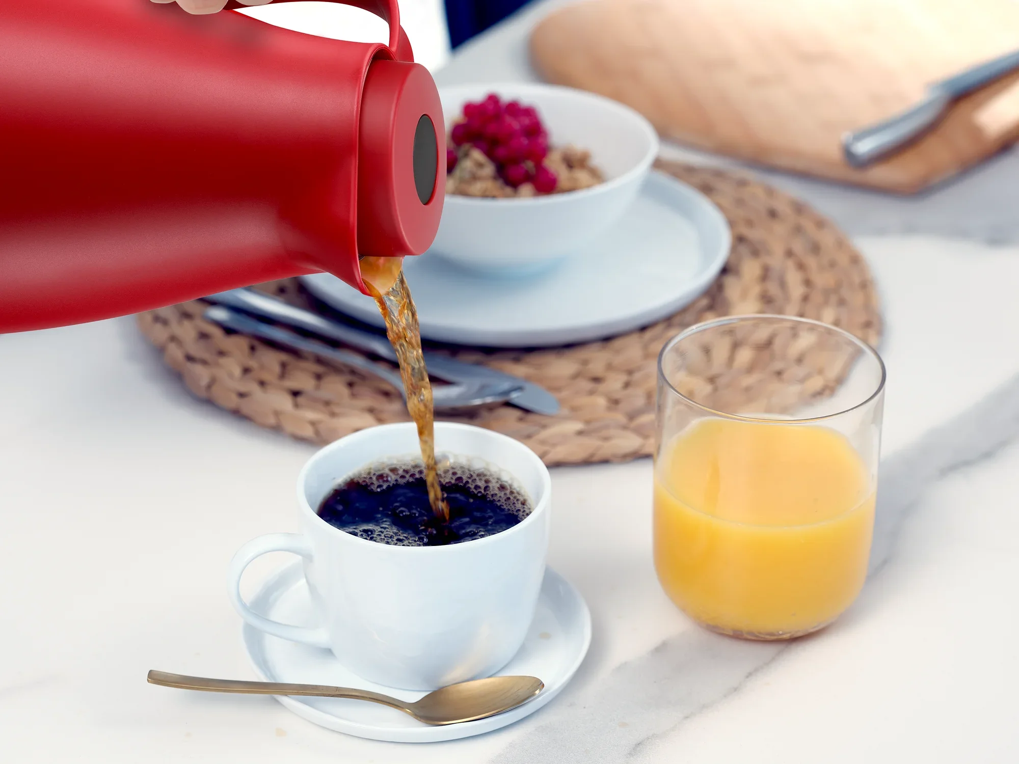 Een rode isoleerkan genaamd Harmonic schenkt hete koffie in een wit kopje. Ernaast staat een glas sinaasappelsap op een tafel gedecoreerd met een geweven placemat.