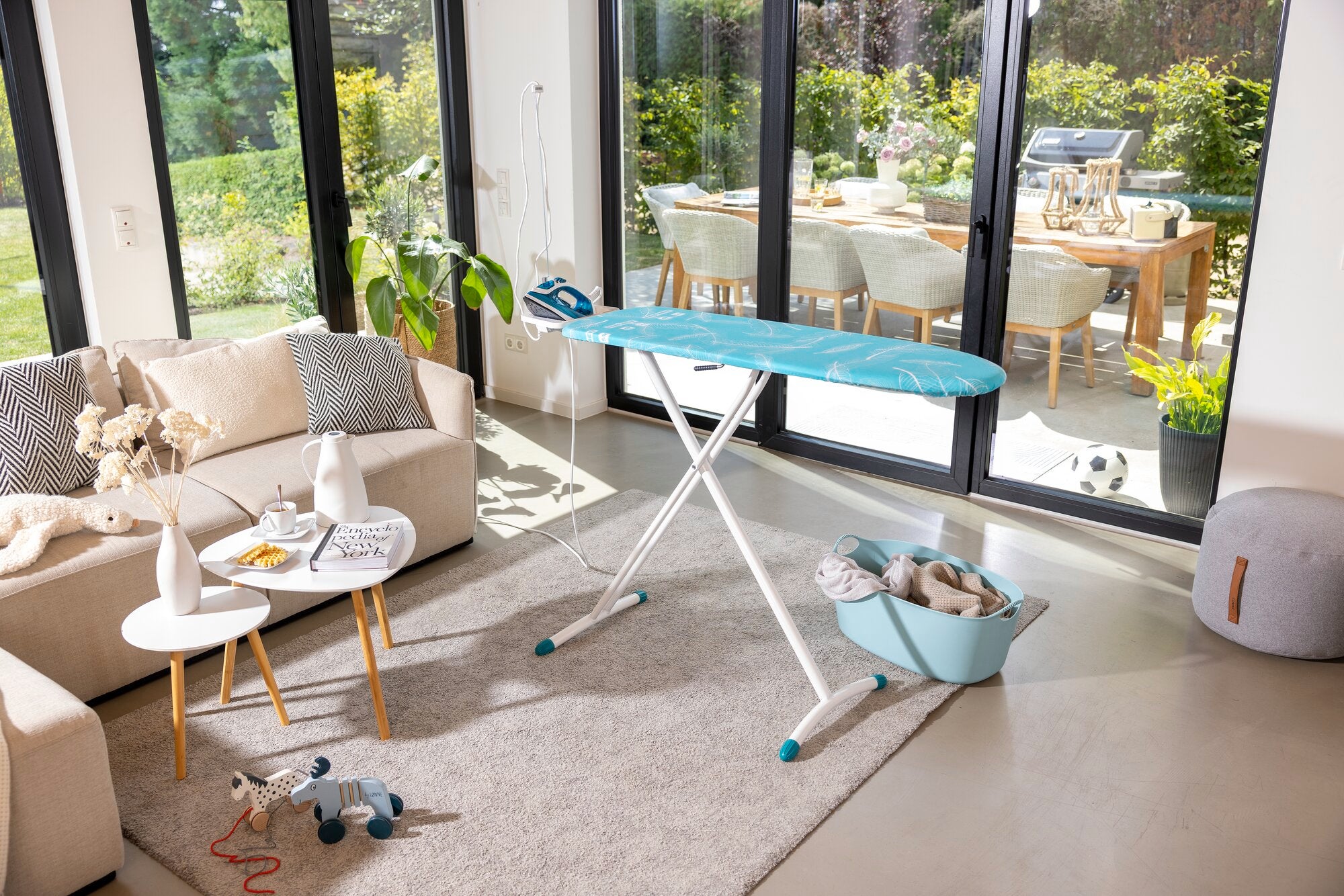 Bügelbrett Leifheit Air Board M Solid in einem modernen Wohnzimmer. Es steht auf einem Teppich vor großen Fenstern mit Blick auf einen sonnigen Garten.