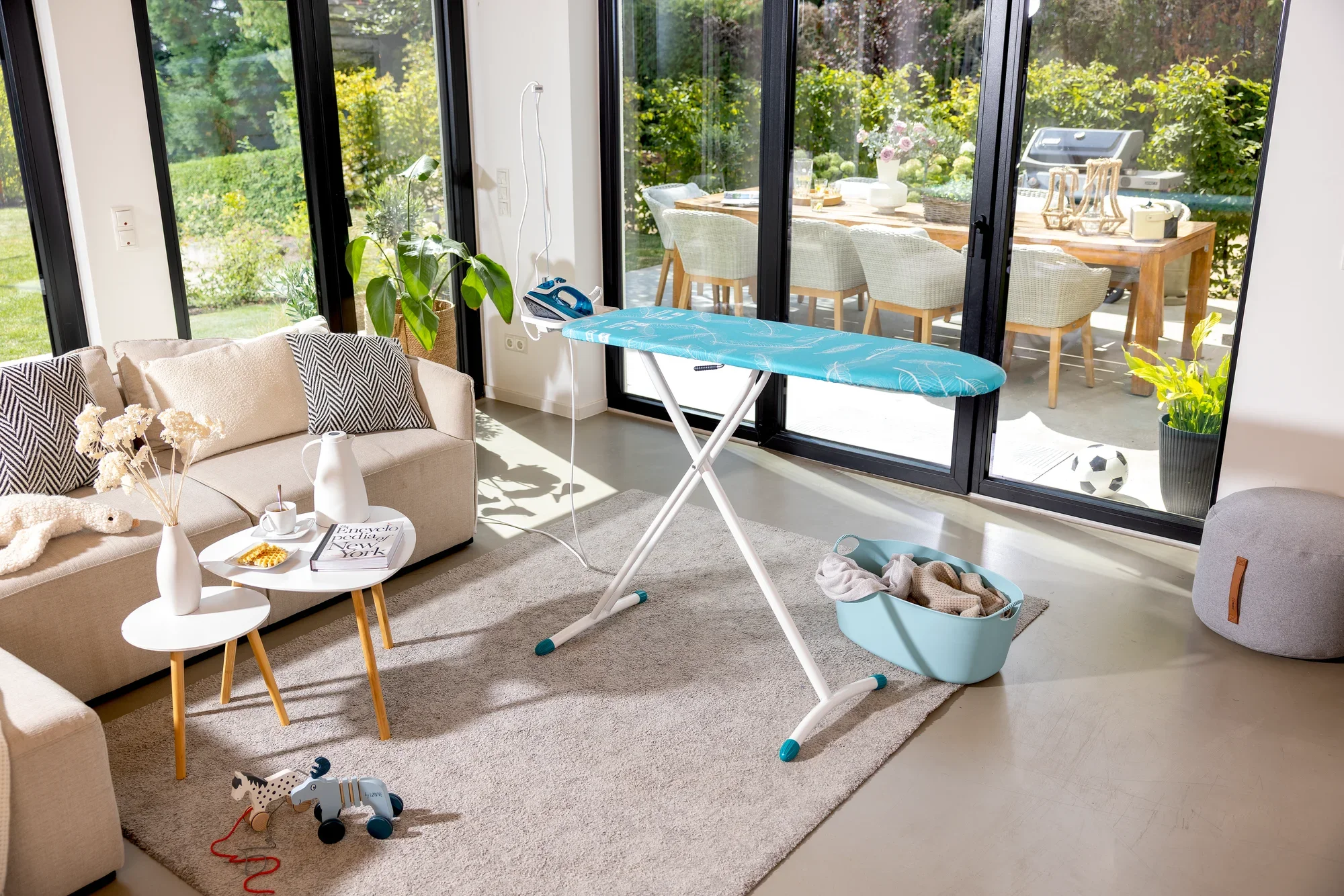 Bügelbrett Leifheit Air Board M Solid in einem modernen Wohnzimmer. Es steht auf einem Teppich vor großen Fenstern mit Blick auf einen sonnigen Garten.
