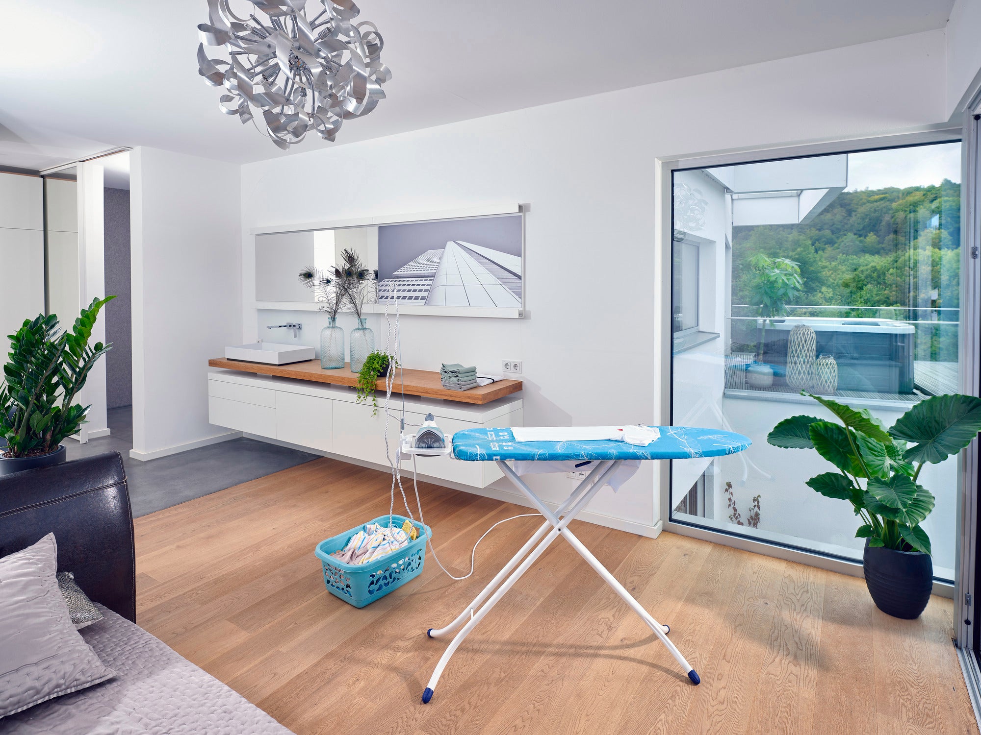 Tabla de planchar Air Board M Compact Plus en una habitación moderna y luminosa con grandes ventanas. La tabla se encuentra sobre un suelo de madera, rodeada de plantas y un cesto para la ropa.
