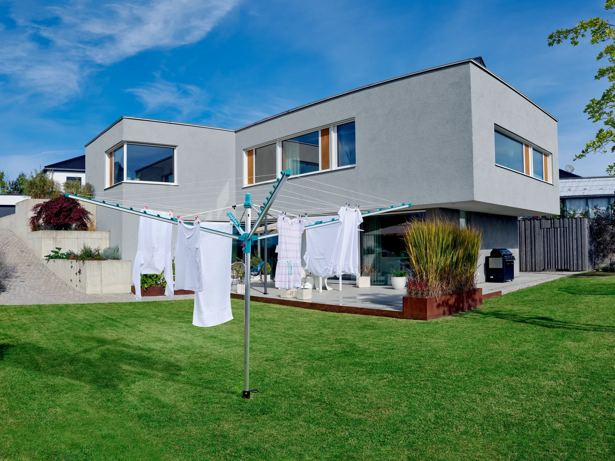 Nowoczesny parasol na pranie Linomatic 500 Easy stoi w zadbanym ogrodzie przed nowoczesnym domem. Białe pranie wisi na linkach do wyschnięcia.