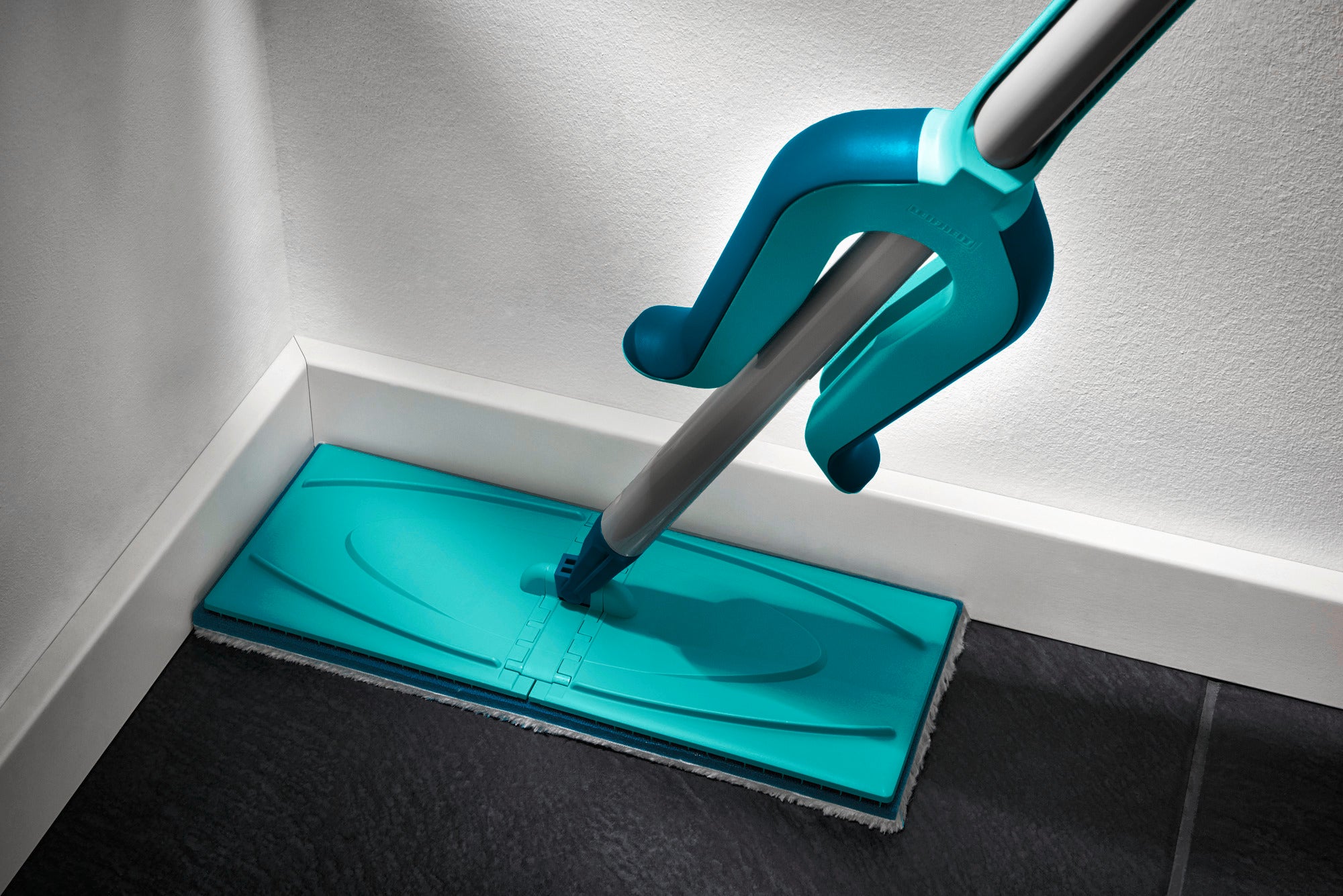 Ein türkisfarbener Bodenwischer, Modell Picobello XL, reinigt eine Ecke eines dunklen Fliesenbodens. Der Wischer hat ein flaches, rechteckiges Design für effiziente Reinigung.