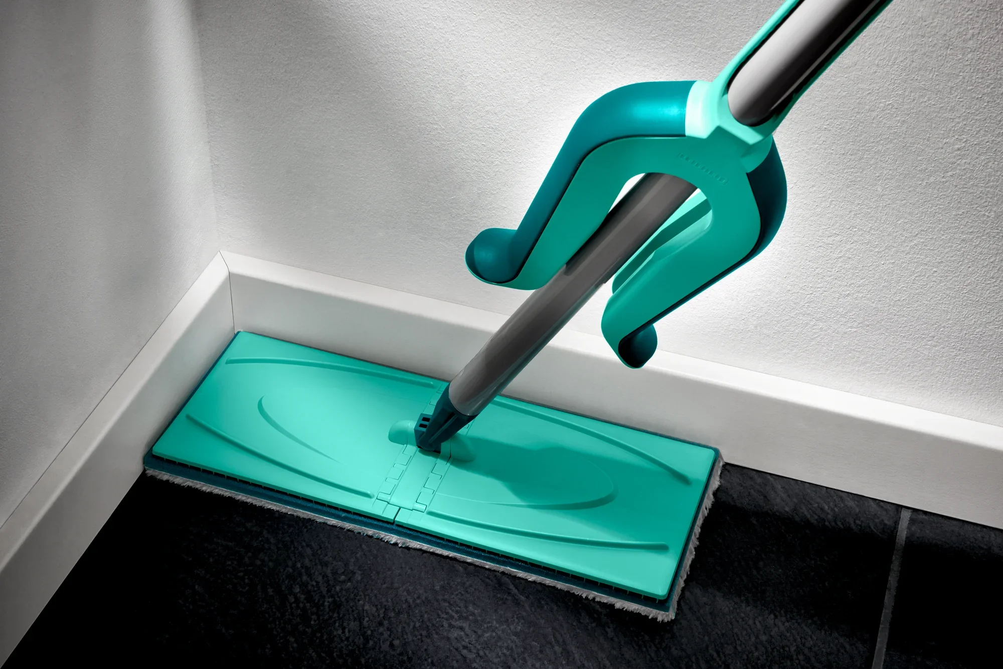 Una mopa turquesa, modelo Picobello XL, limpia una esquina de un suelo de baldosas oscuras. La mopa tiene un diseño plano y rectangular para una limpieza eficaz.