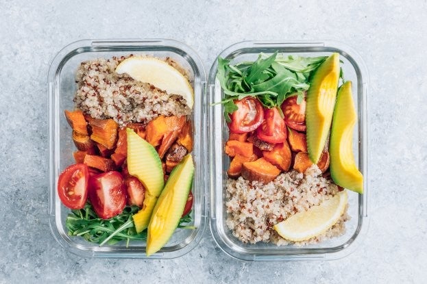 Zwei Glasbehaelter mit Quinoa, Avocado, Tomaten, Suesskartoffeln, Rucola und Zitronenscheiben auf grauem Untergrund.