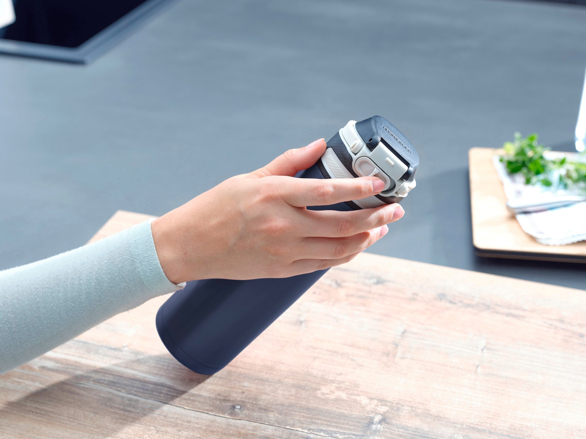 Una mano tiene un thermos Flip blu scuro con una capacità di 600 ml. Il thermos ha una chiusura argentata ed è appoggiato su un tavolo di legno. Sullo sfondo si vedono utensili da cucina.