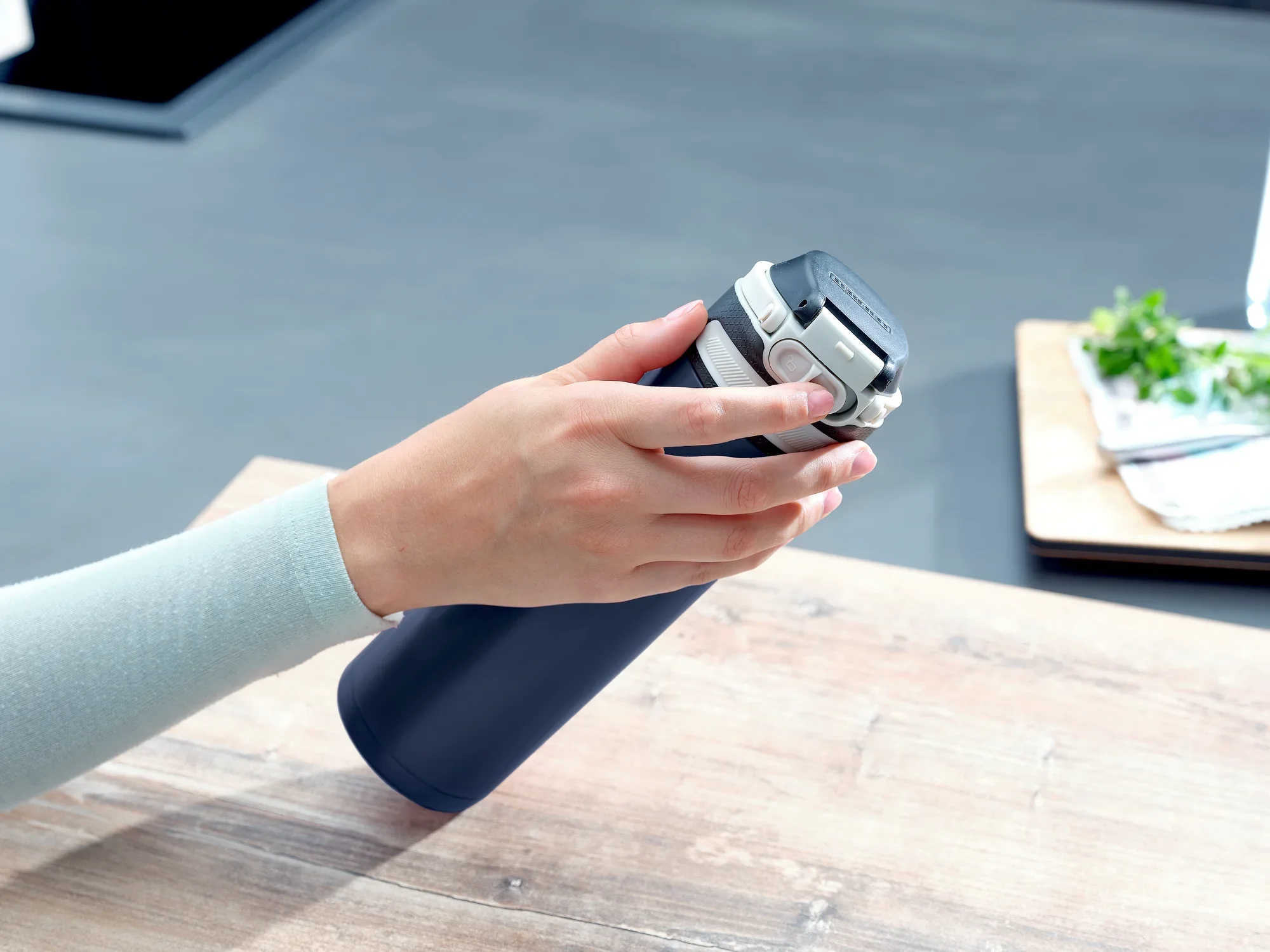 Una mano sostiene un vaso térmico Flip azul oscuro con una capacidad de 600 ml. El vaso tiene un cierre plateado y está sobre una mesa de madera. Al fondo se ven utensilios de cocina.