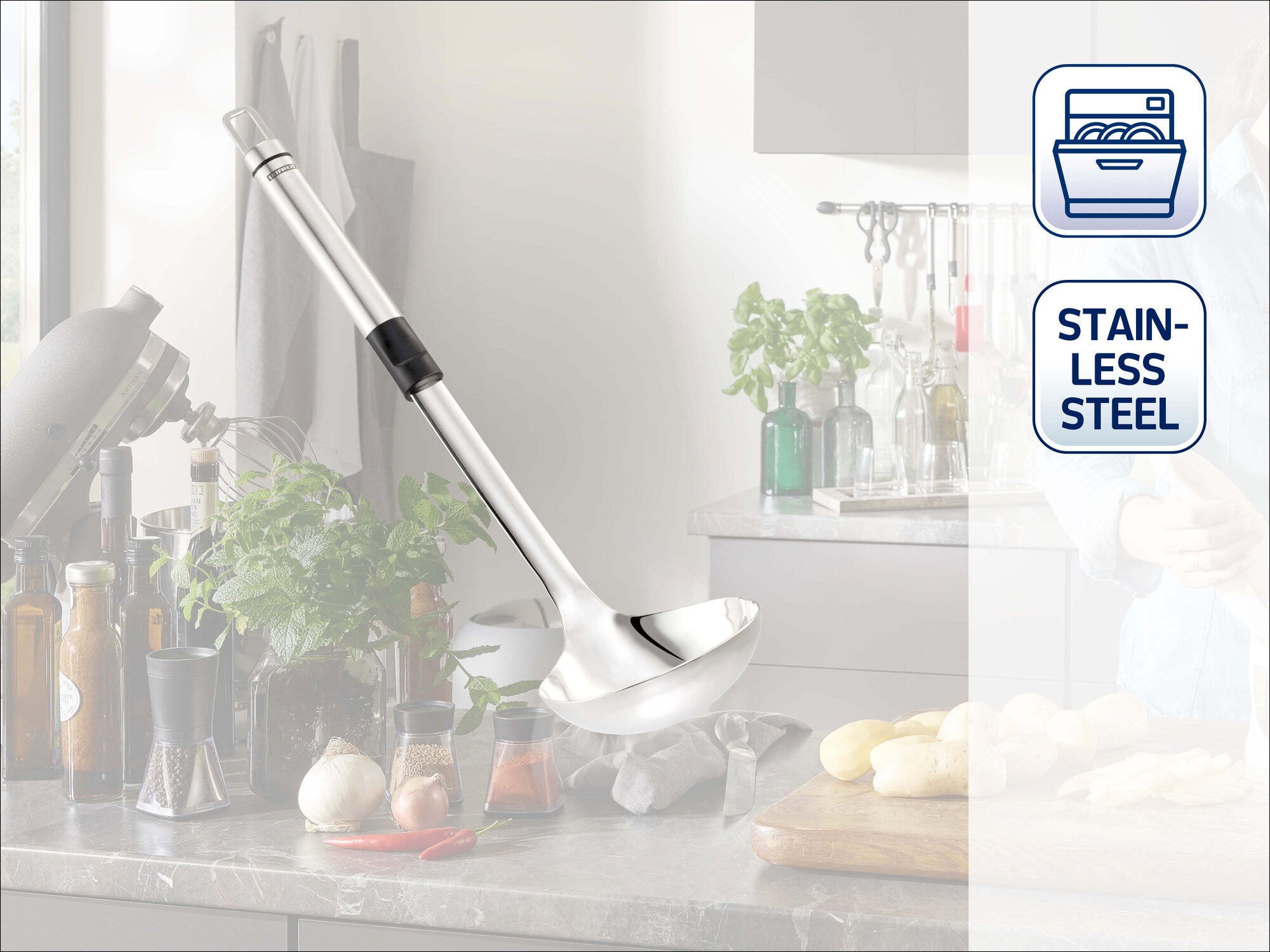 Leifheit Schöpflöffel aus Edelstahl mit langem Griff und schwarzem Soft-Touch-Element, dekorativ an einer modernen Küchenwand hängend, vor heller Küchenkulisse mit Kräutern, Flaschen und Schneidebrett.