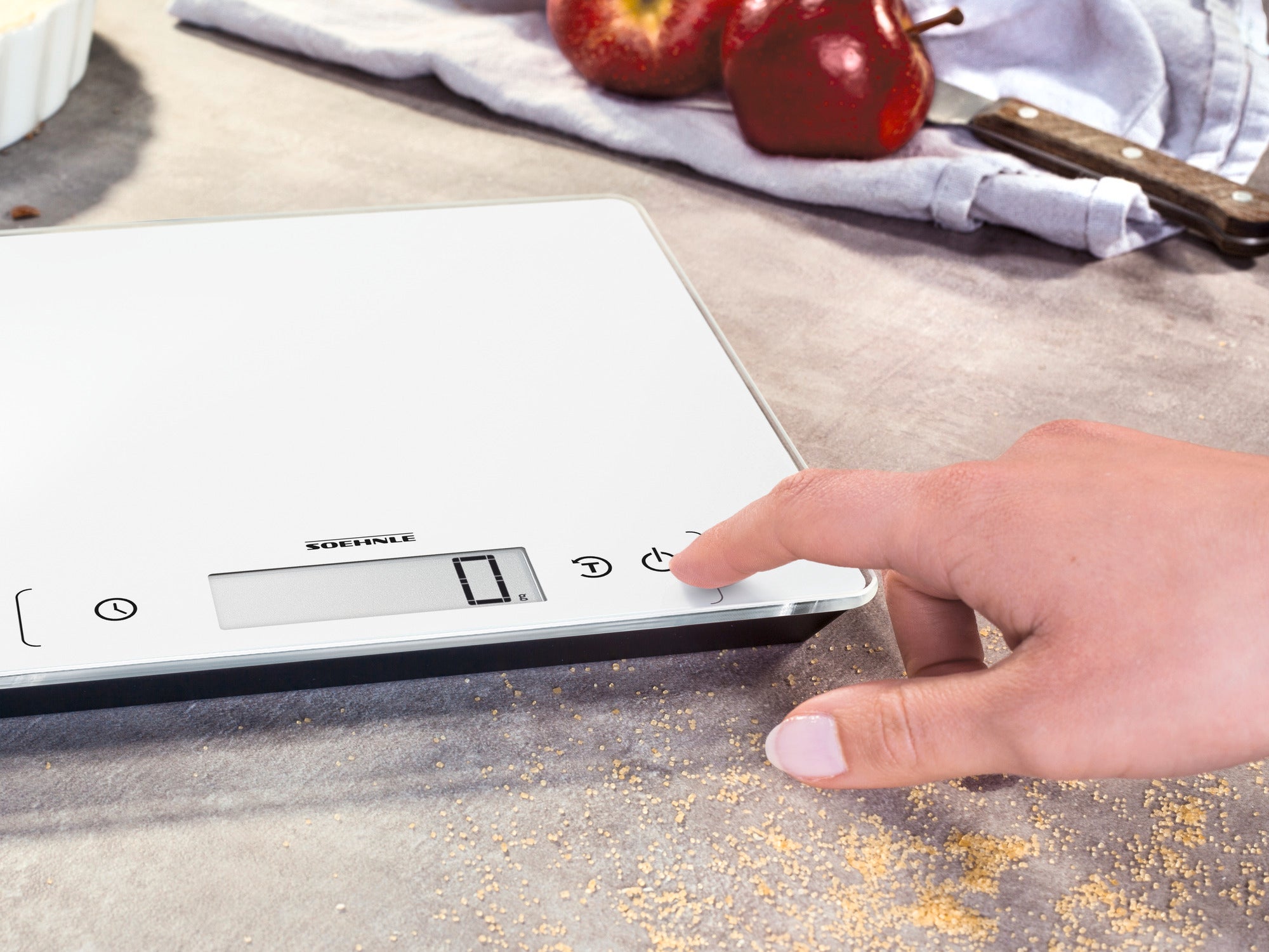 Eine digitale Küchenwaage, Modell Page Comfort 400, wird auf einer Küchenarbeitsplatte verwendet. Eine Hand bedient die Waage, während im Hintergrund Äpfel und ein Handtuch zu sehen sind.
