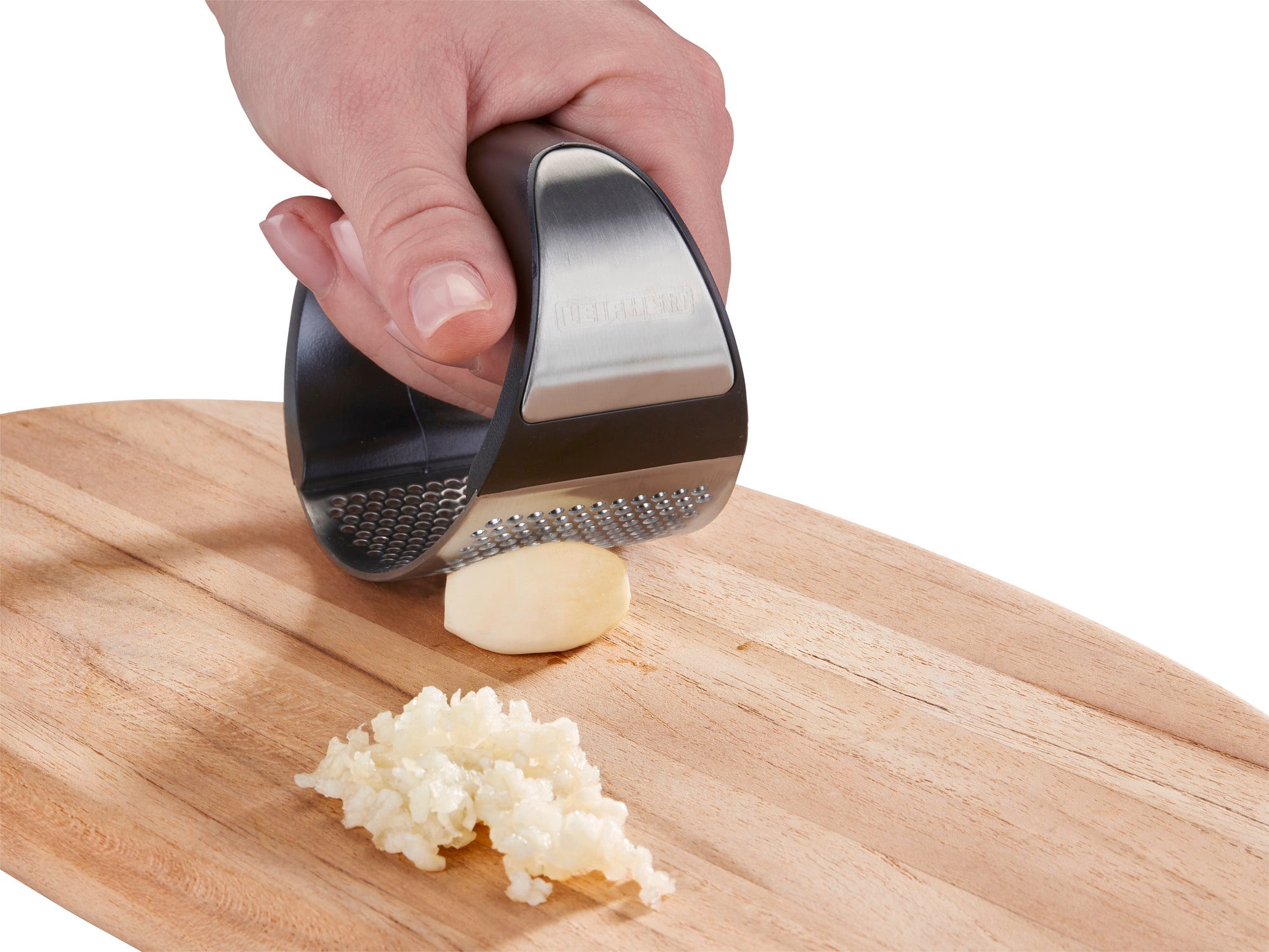 Eine Hand benutzt die Knoblauchpresse "Knobi King", um eine Knoblauchzehe auf einem Holzbrett zu zerdrücken. Der gepresste Knoblauch liegt fein zerkleinert daneben.