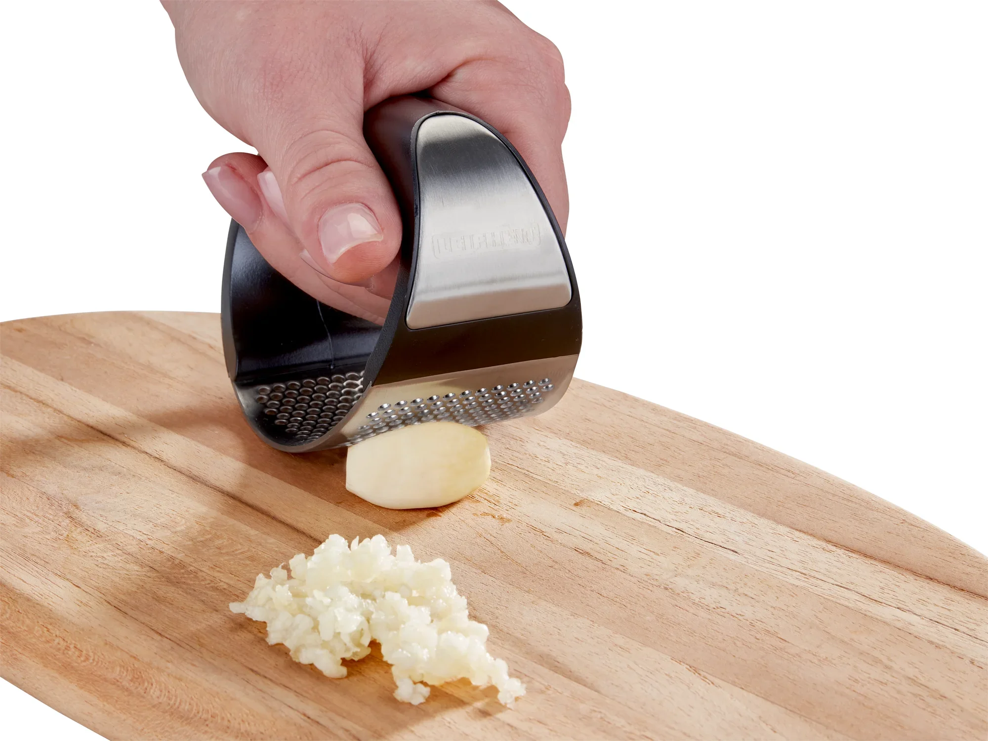 Une main utilise le presse-ail « Knobi King » pour écraser une gousse d'ail sur une planche en bois. L'ail pressé est finement haché à côté.