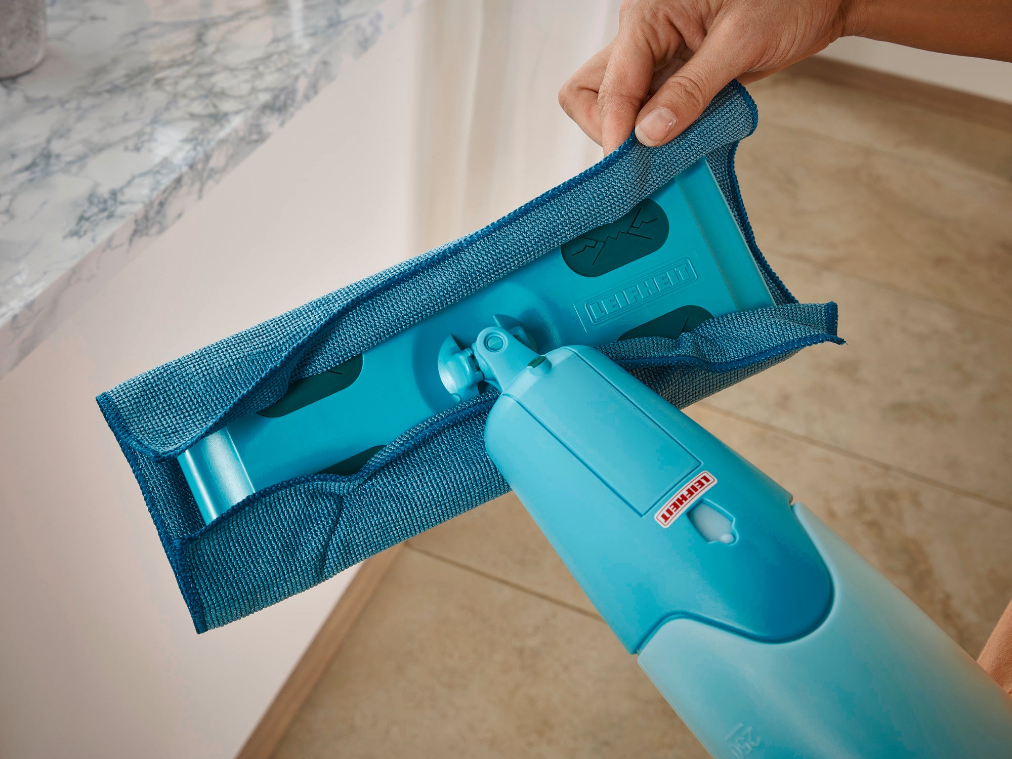 Een blauwe spraymop Pico Spray wordt gebruikt op een lichtgekleurde tegelvloer. Eén hand houdt de wisser vast terwijl het deksel wordt verwisseld. Ideaal voor snel schoonmaken.