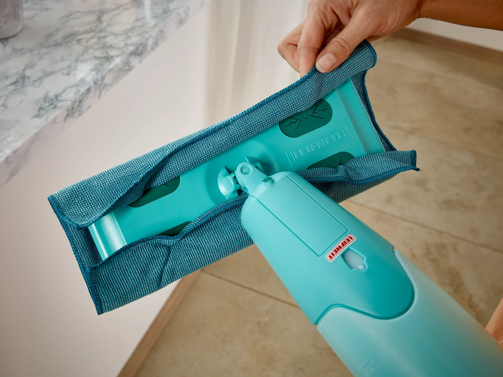 Un balai à franges Pico Spray bleu est utilisé sur un sol carrelé clair. Une main tient le balai pendant que la housse est changée. Idéal pour un nettoyage rapide.