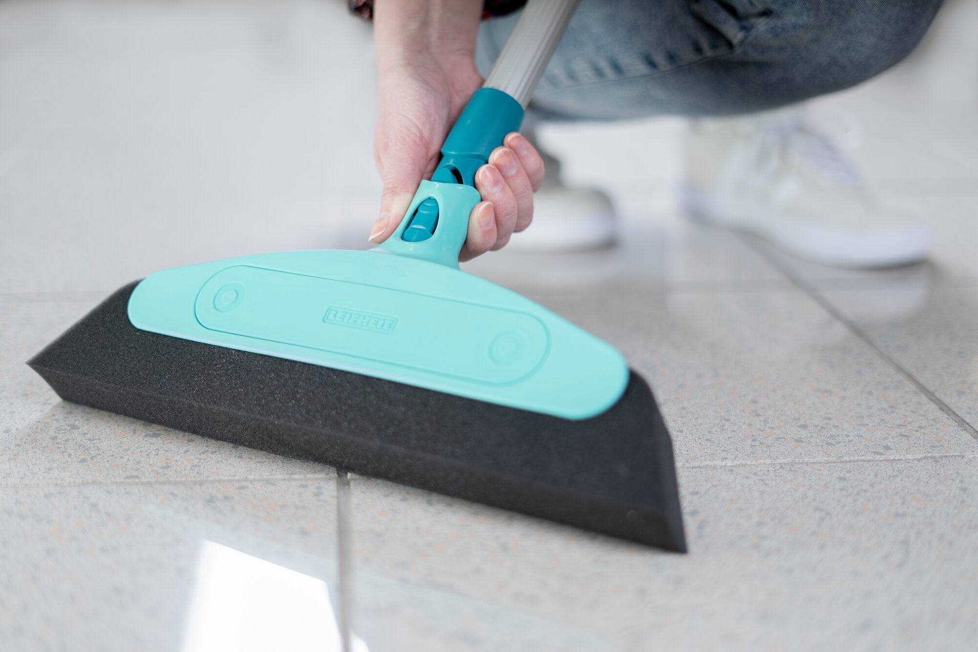 Schuimrubberen bezem in gebruik op een lichtgekleurde tegelvloer. De bezem heeft een turquoise kliksysteem en wordt met één hand vastgehouden. Ideaal voor gladde oppervlakken.