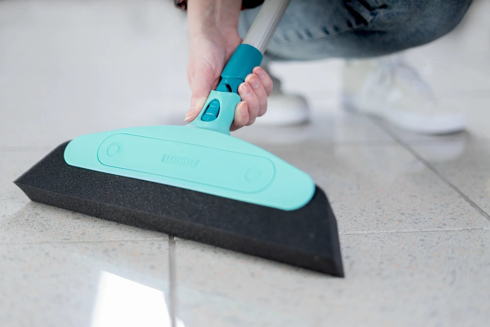 Balai en mousse utilisé sur un sol carrelé clair. Le balai est doté d'un système de clic turquoise et se tient d'une seule main. Idéal pour les surfaces lisses.