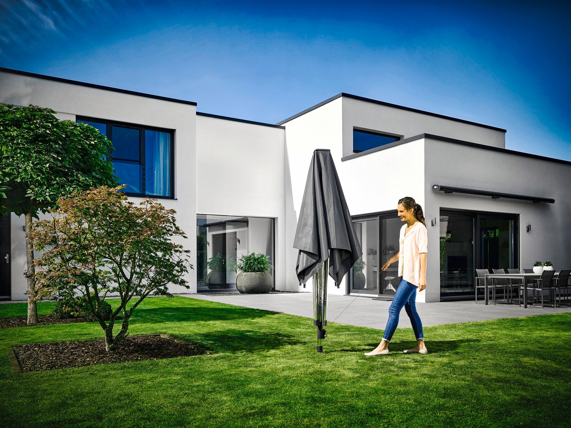 Eine Frau geht im Garten eines modernen Hauses an einer geschlossenen Leifheit LinoProtect 400 Wäschespinne vorbei. Der Himmel ist klar und die Umgebung wirkt gepflegt und einladend.