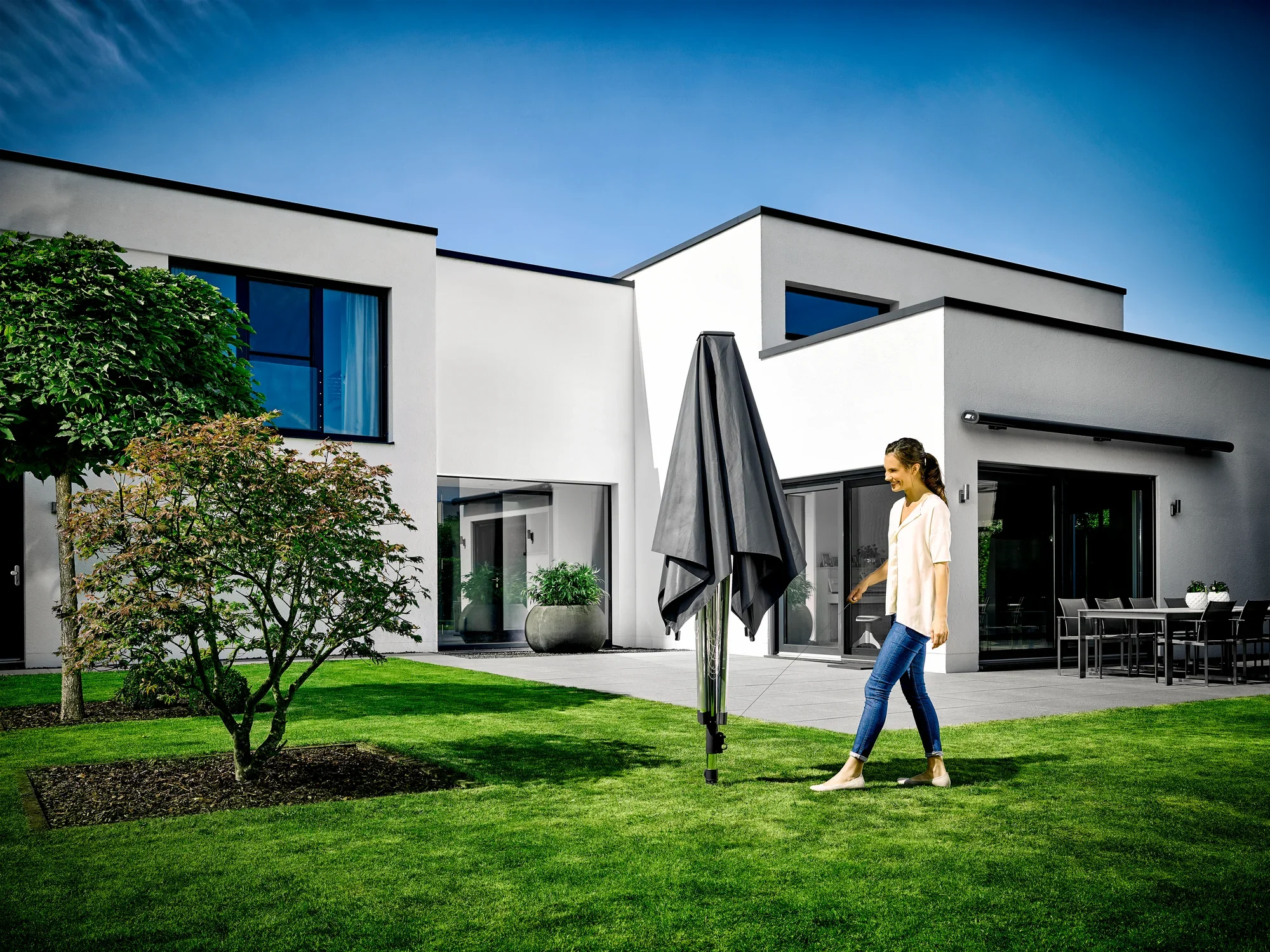 Une femme passe devant un séchoir à linge Leifheit LinoProtect 400 fermé dans le jardin d'une maison moderne. Le ciel est dégagé et l'environnement semble soigné et accueillant.