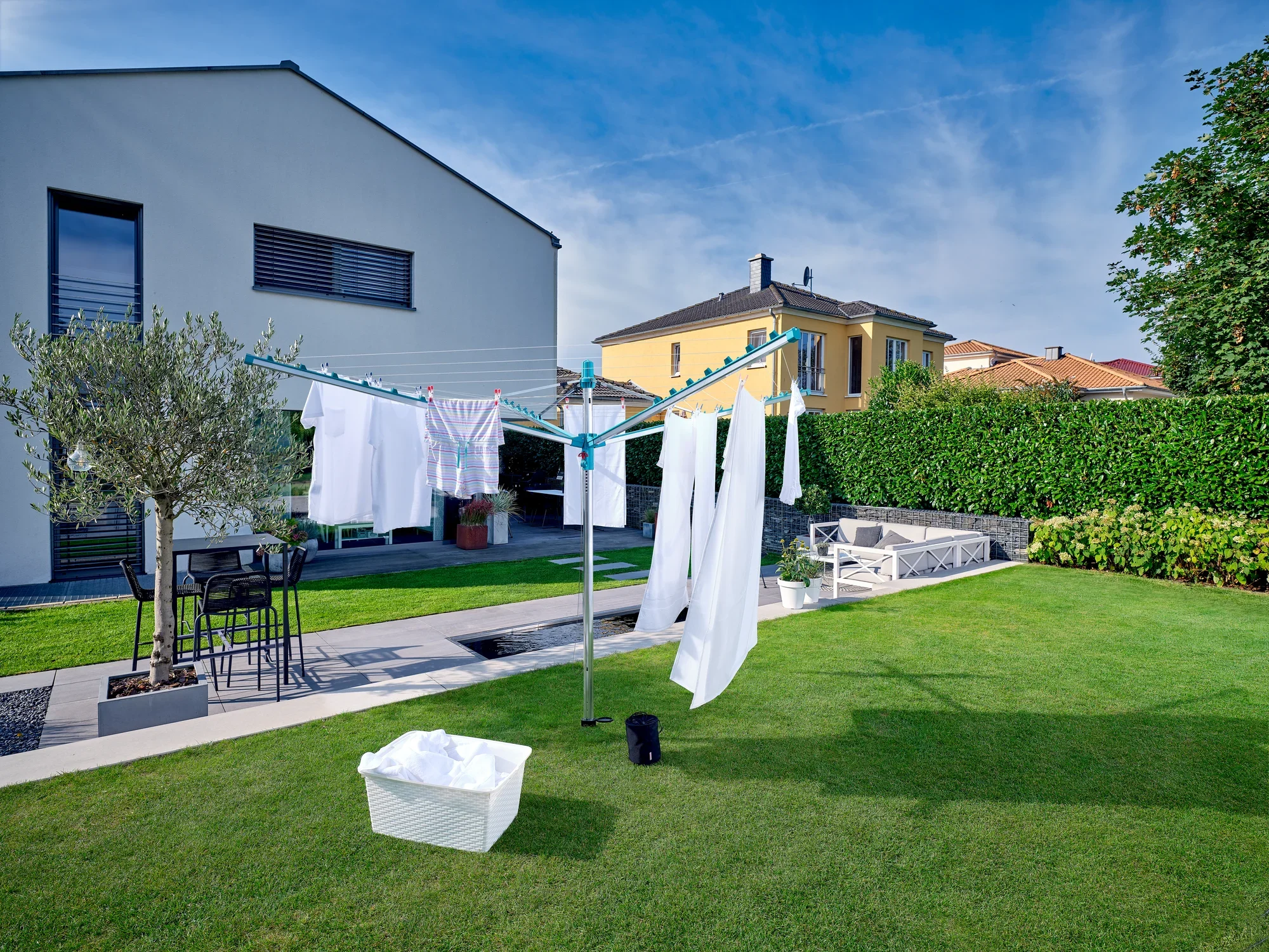 Parasol na pranie Linomatic 400 Easy stoi w zadbanym ogrodzie. Świeże pranie wisi na linkach do wyschnięcia. W tle widać nowoczesne domy i zielony trawnik.