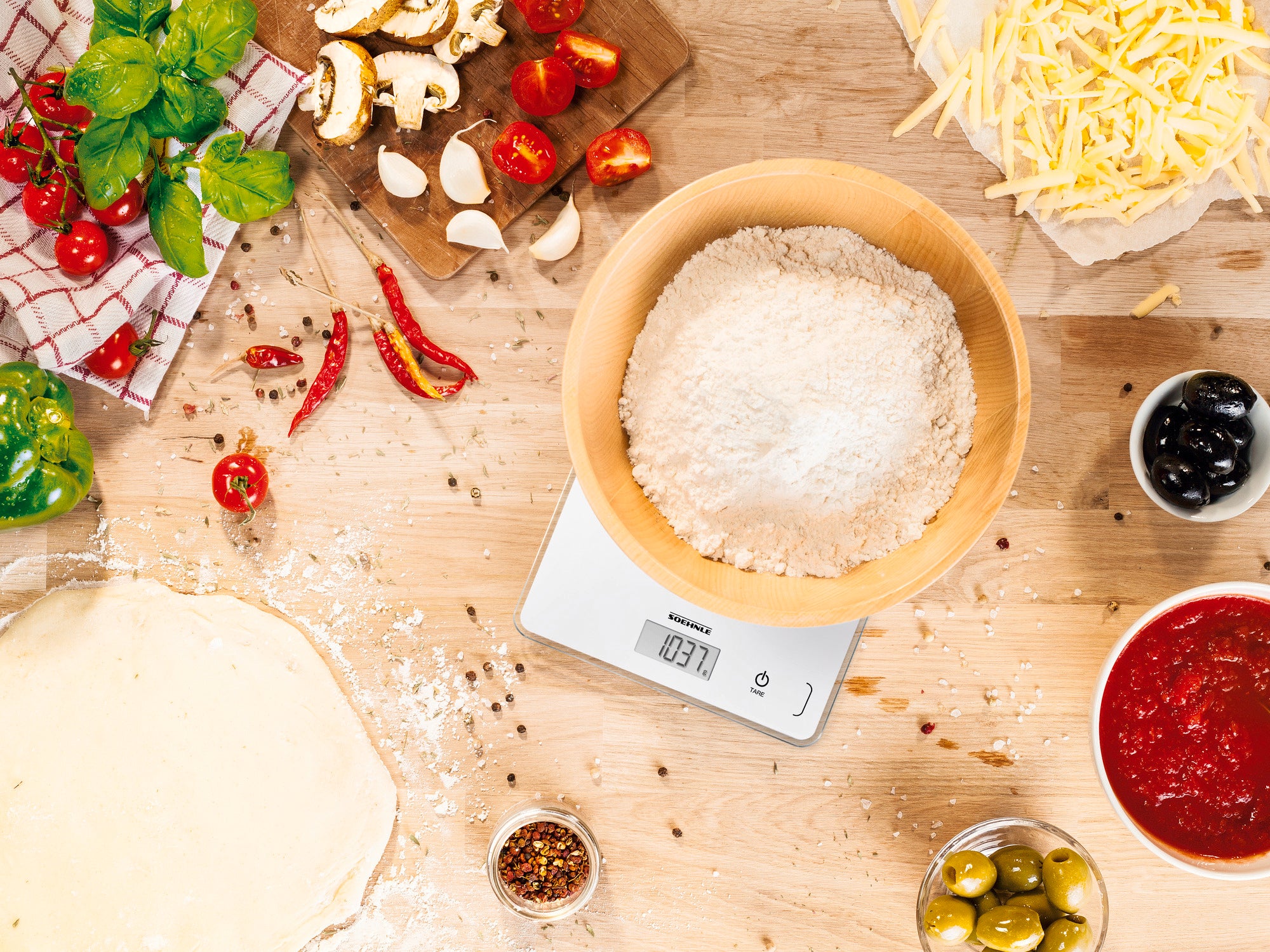 Digitale keukenweegschaal Page Compact 300 in actie. Een kom bloem staat op de weegschaal, omringd door ingrediënten zoals tomaten, knoflook en olijven. Ideaal om te bakken.