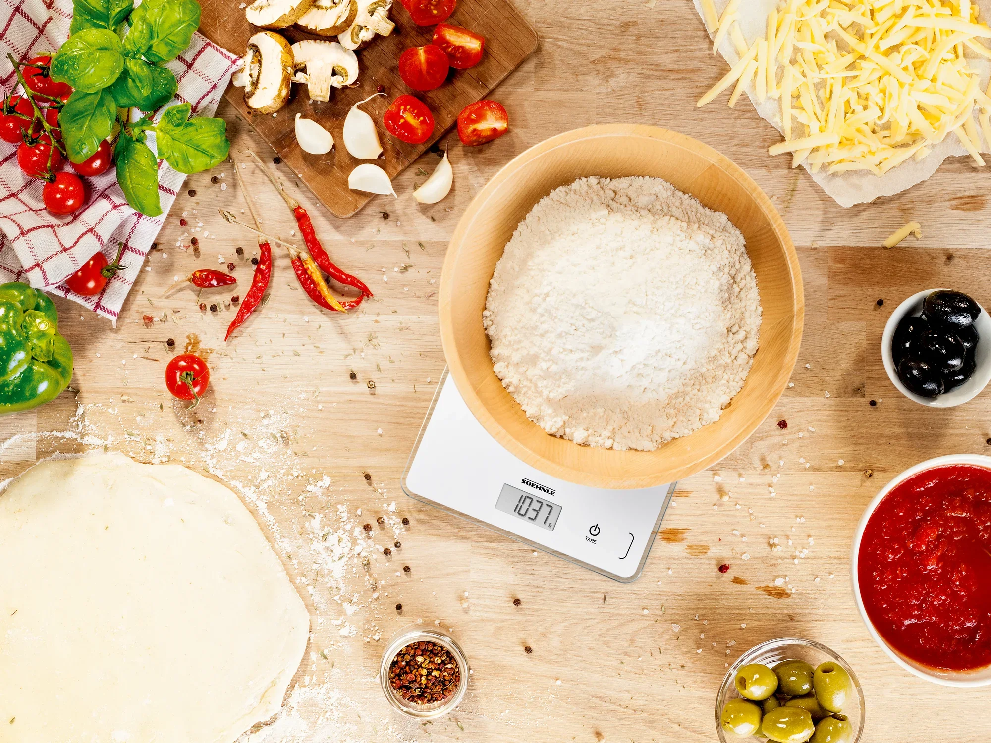 Page Compact 300 digital kitchen scale in action. A bowl of flour stands on the scale, surrounded by ingredients such as tomatoes, garlic and olives. Ideal for baking.