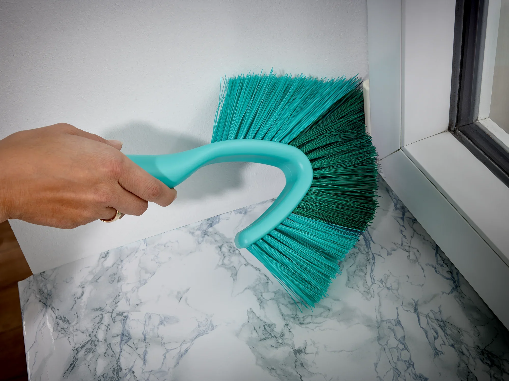 Een hand houdt de SUPERDUSTER bezem met telescopische steel vast om stof te verwijderen van een marmeren oppervlak. De bezem heeft blauwe haren en een ergonomisch handvat.