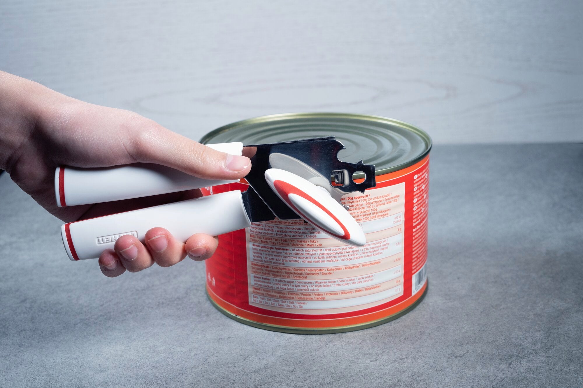 Une main utilise un ouvre-boite blanc et rouge pour ouvrir une grande boite de conserve rouge posee sur une surface grise.
