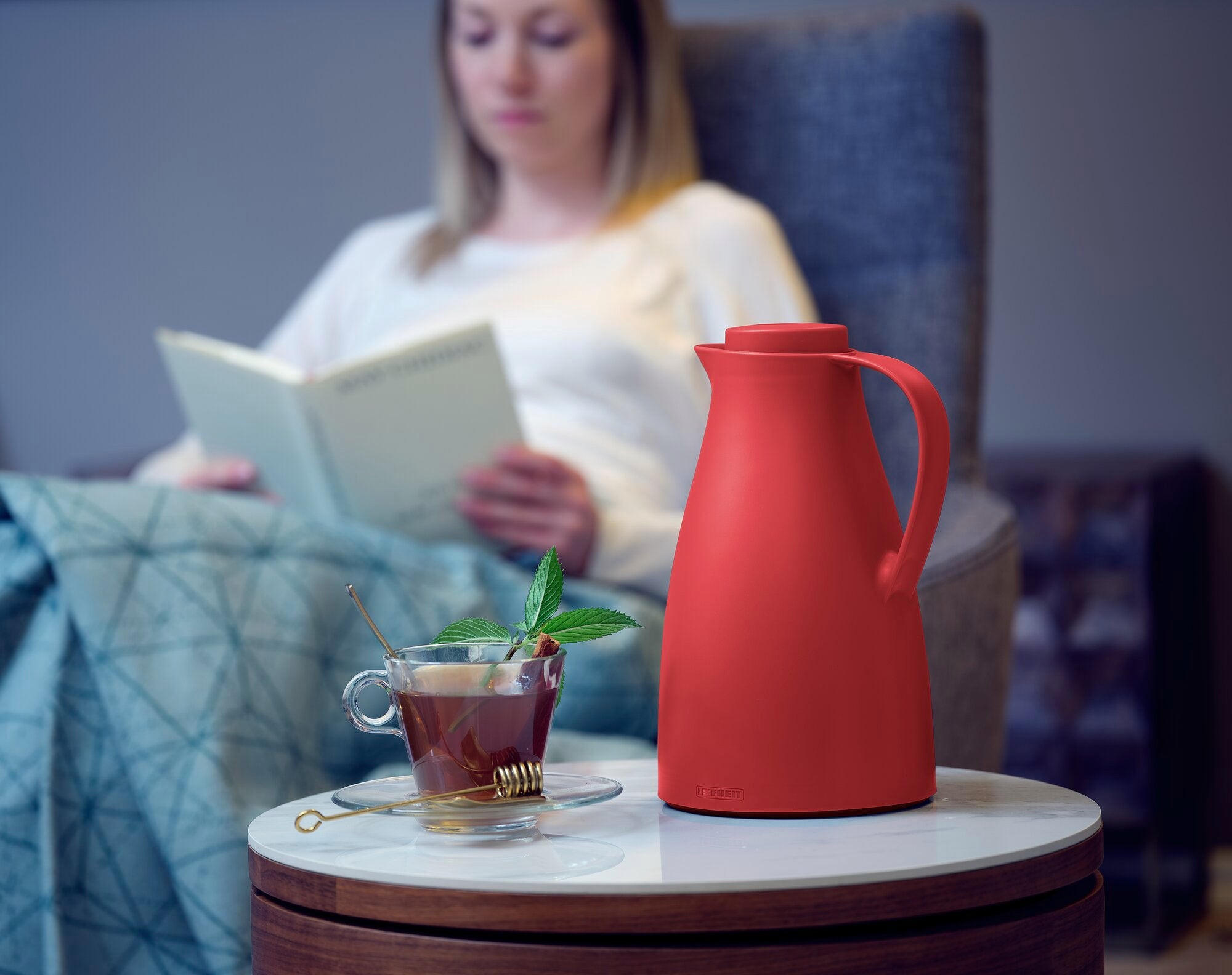 Isolierkanne Harmonic in Rot auf einem Tisch neben einer Tasse Tee mit Minzblättern. Im Hintergrund sitzt eine Person auf einem Sessel und liest ein Buch.