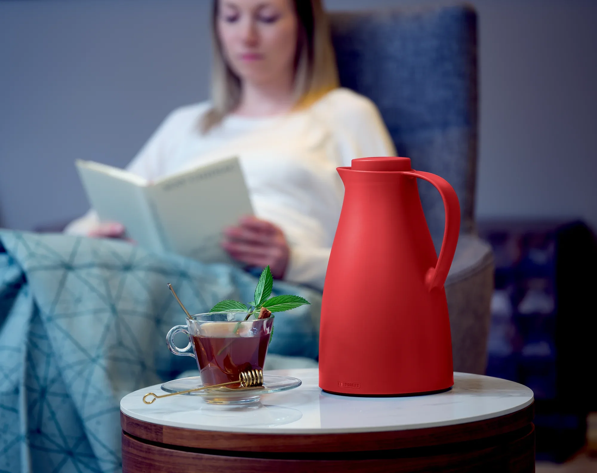 Harmonic isoleerkan in rood op een tafel naast een kopje thee met muntblaadjes. Op de achtergrond zit een persoon in een leunstoel en leest een boek.