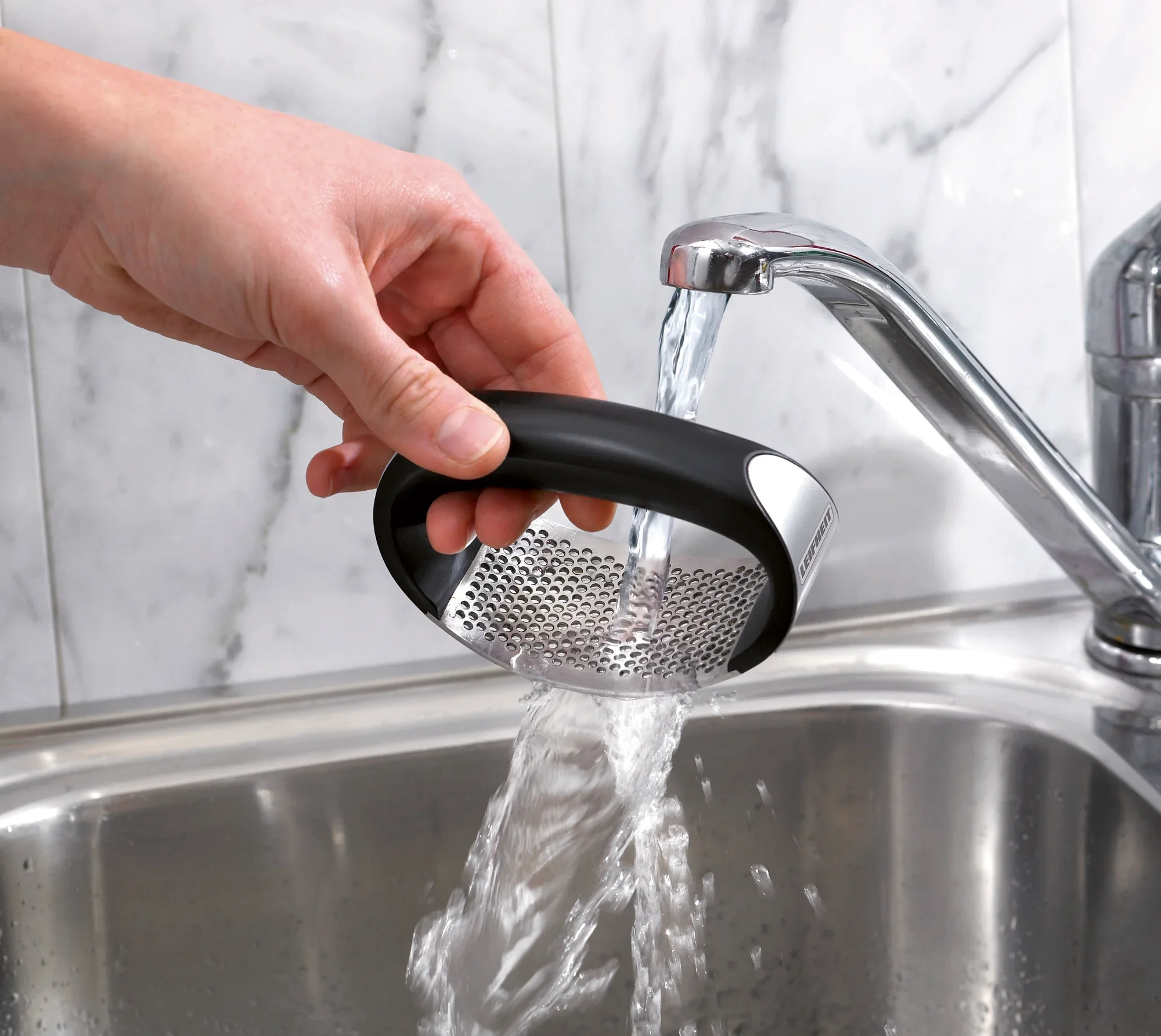 Une main tient le presse-ail « Knobi King » sous l'eau courante dans un évier en acier inoxydable. Le presse-ail est doté d'une poignée ergonomique et d'une grille métallique robuste. Idéal pour un nettoyage facile après utilisation.