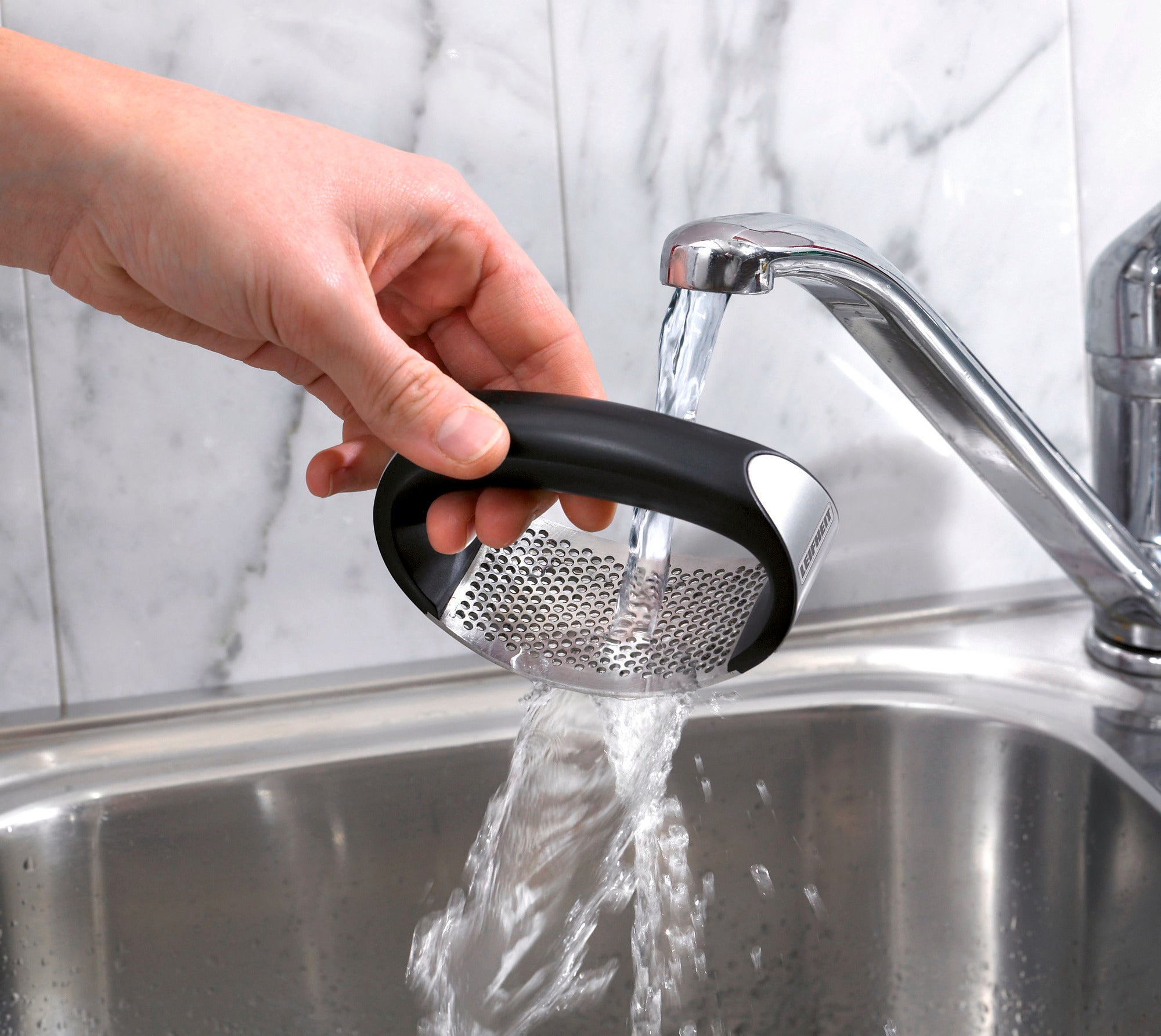 One hand holds the Knobi King garlic press under running water in a stainless steel sink. The press has an ergonomic handle and a sturdy metal grid. Ideal for easy cleaning after use.