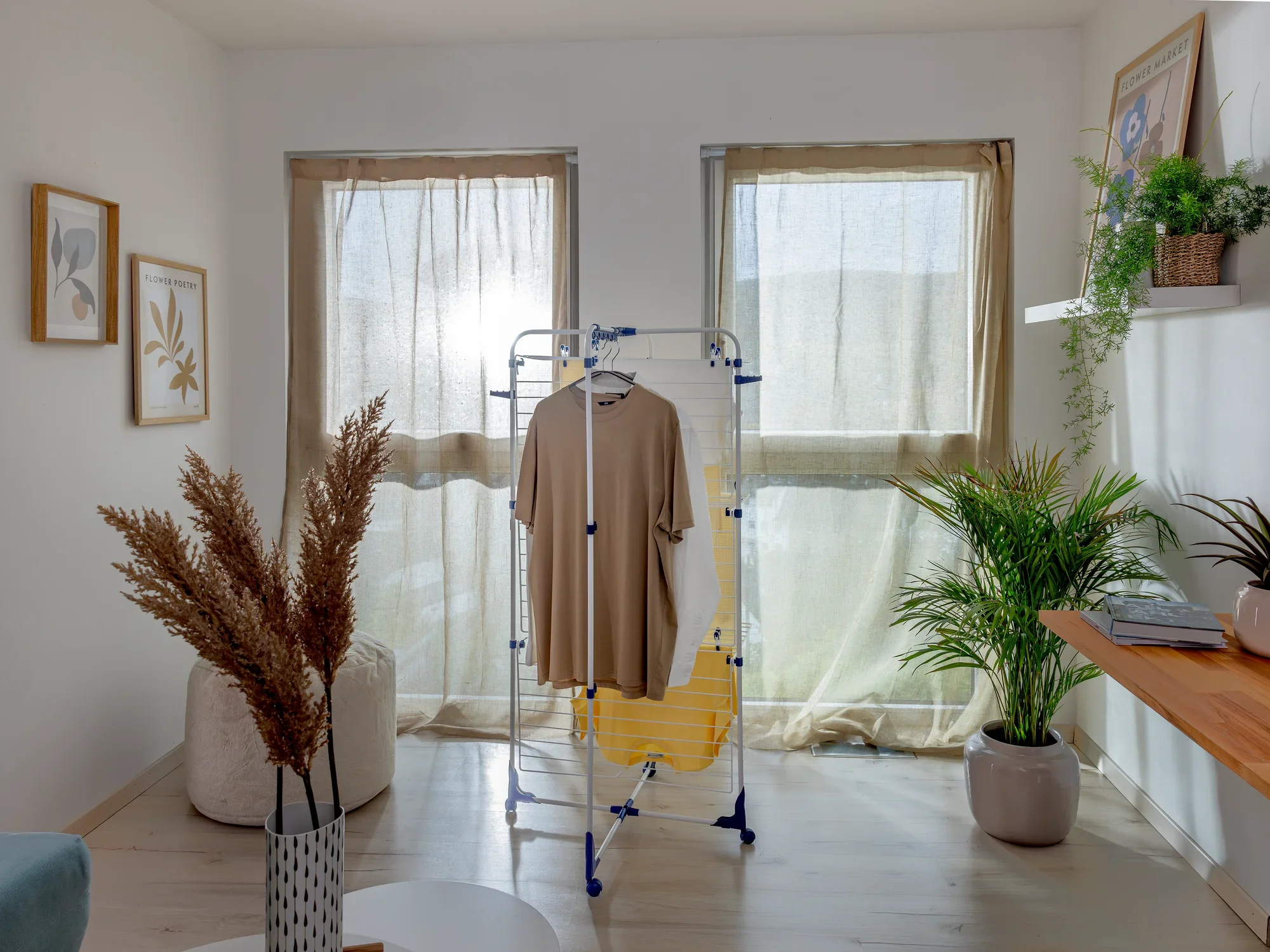 Een Classic Tower 340 toren droogtoren staat in een lichte kamer voor twee ramen. Er hangt een beige kledingstuk aan. Planten en decoraties omringen de wasdroger.
